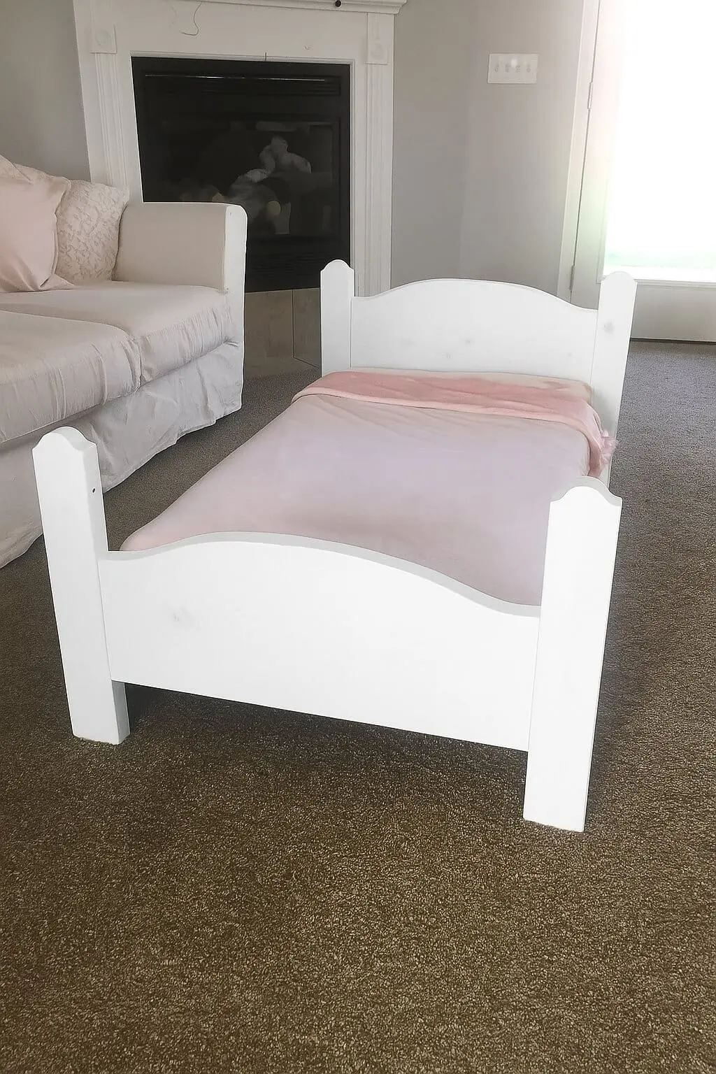 Front view of a white DIY toddler bed with curved headboard and footboard, handmade from leftover wood and styled with pale pink bedding.