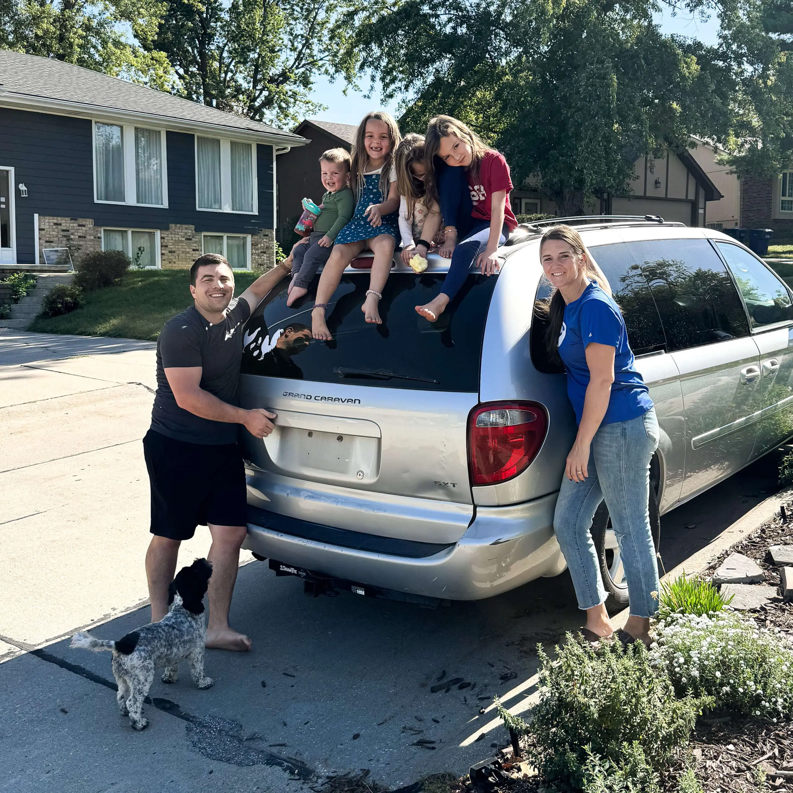 Happy family standing beside their silver minivan before a road trip with kids sitting on the back of the vehicle and a small dog at their feet.