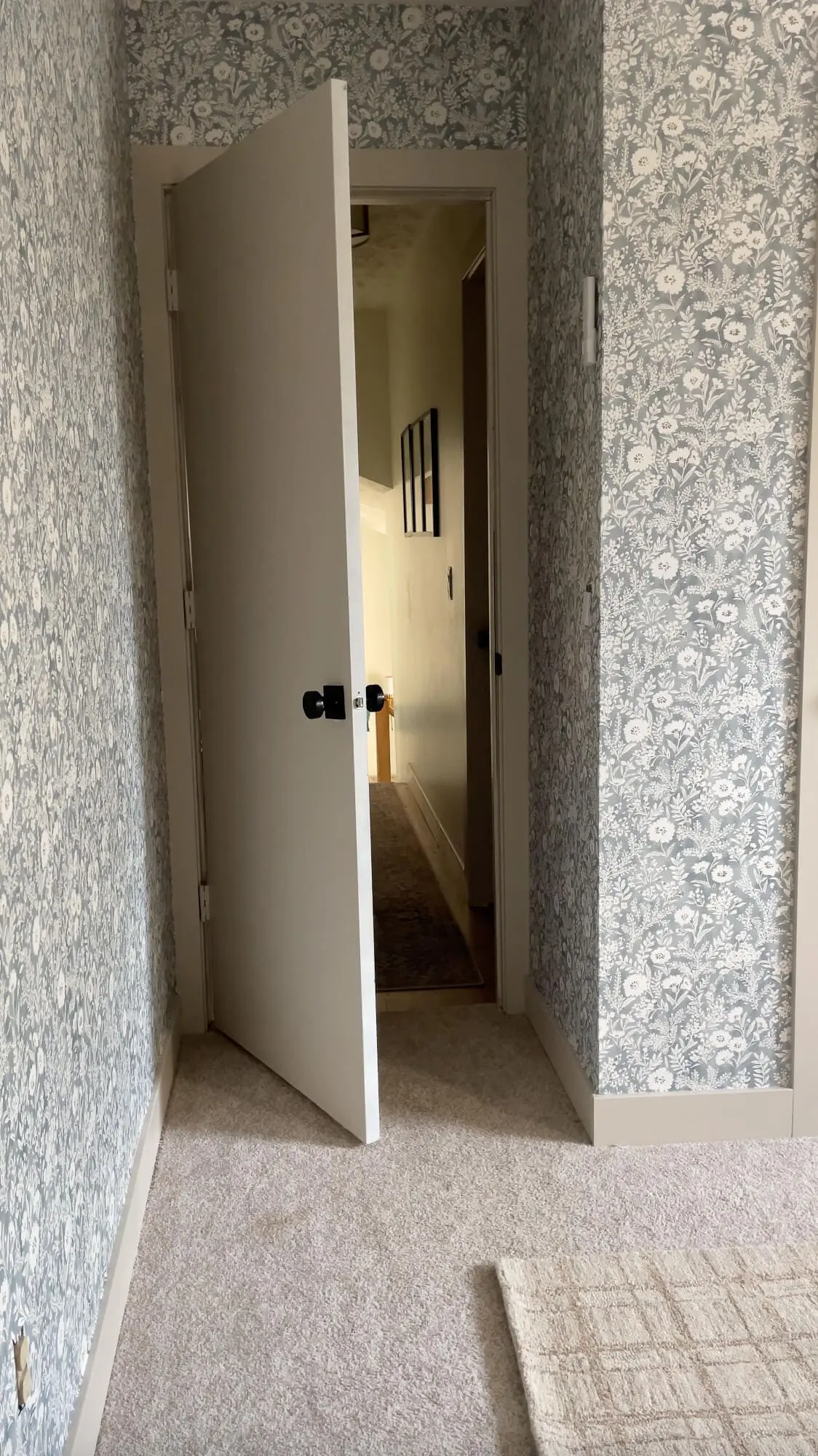 View from a carpeted bedroom into a hallway, with walls fully covered in floral wallpaper and a partially open door.