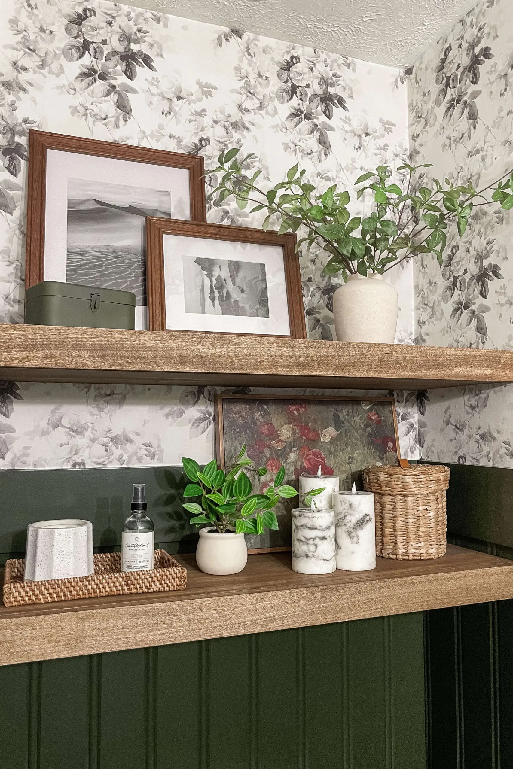 Floating wood shelves styled with greenery, candles, and artwork against a soft black and white floral wallpaper, with dark green vertical wall paneling below for contrast.