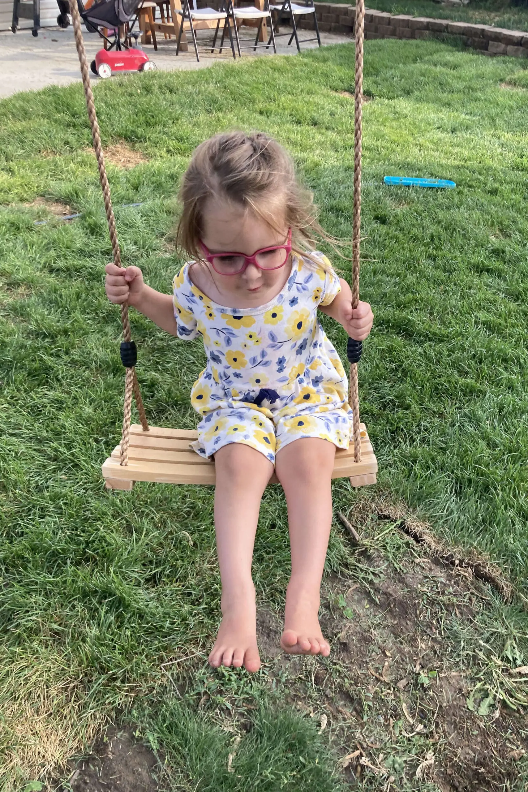 Young girl on a wooden swing with rope handles, concentrating as she balances above a patchy grass area in the backyard.