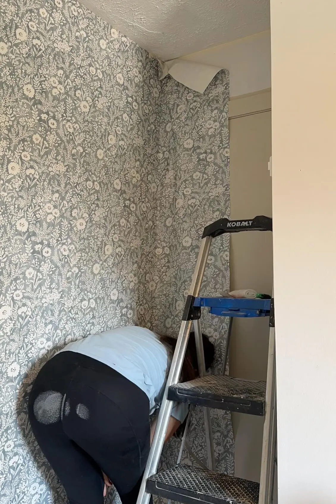 Installing the final wallpaper panel in a tight corner using a ladder, with the floral pattern continuing around the doorway.
