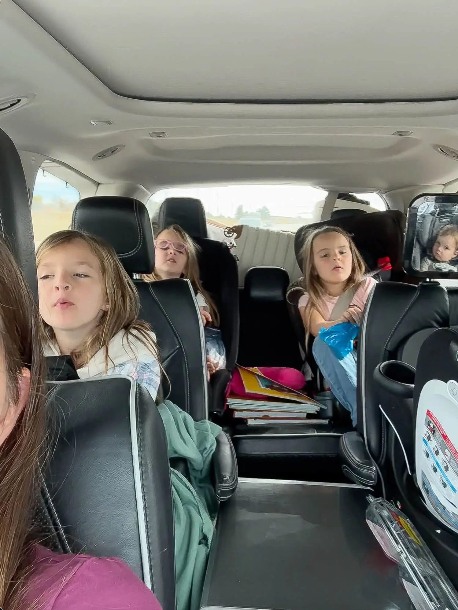 Three kids riding in the backseat of a minivan during a road trip, surrounded by snacks, books, and car seat accessories, with a baby visible in a rear-facing mirror.