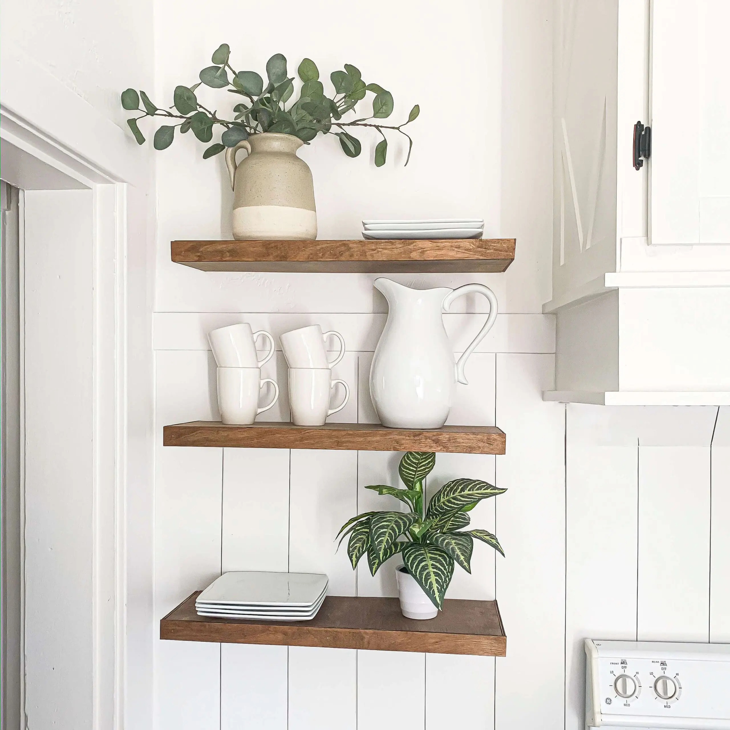 Close-up of three DIY floating shelves in a white shiplap kitchen wall, decorated with white ceramic dishes, a large pitcher, stacked plates, and a leafy potted plant—an affordable and stylish kitchen storage idea.