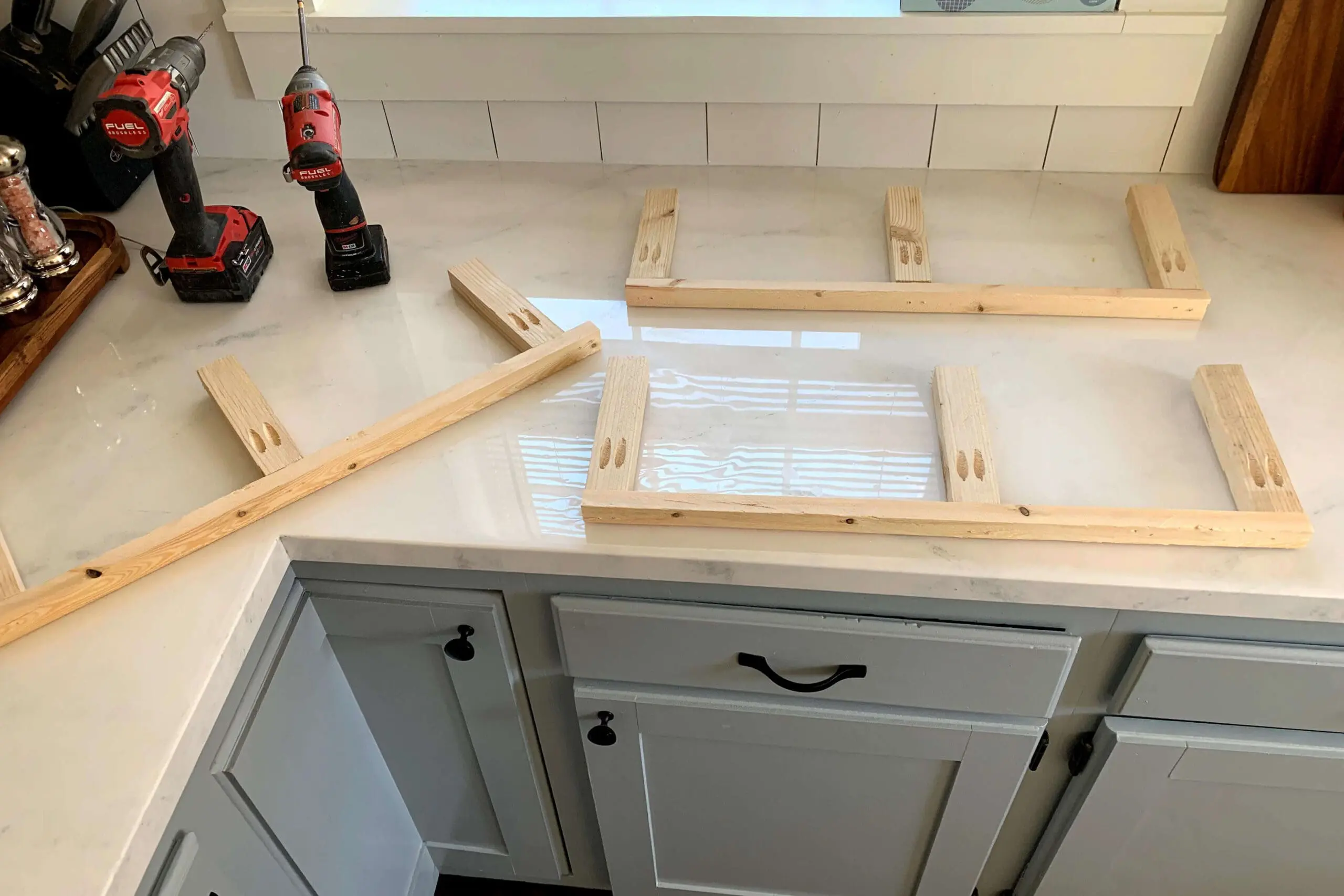 Assembled shelf frames made from wood and pocket hole joinery, laid out on a white kitchen countertop next to power tools during a home project.