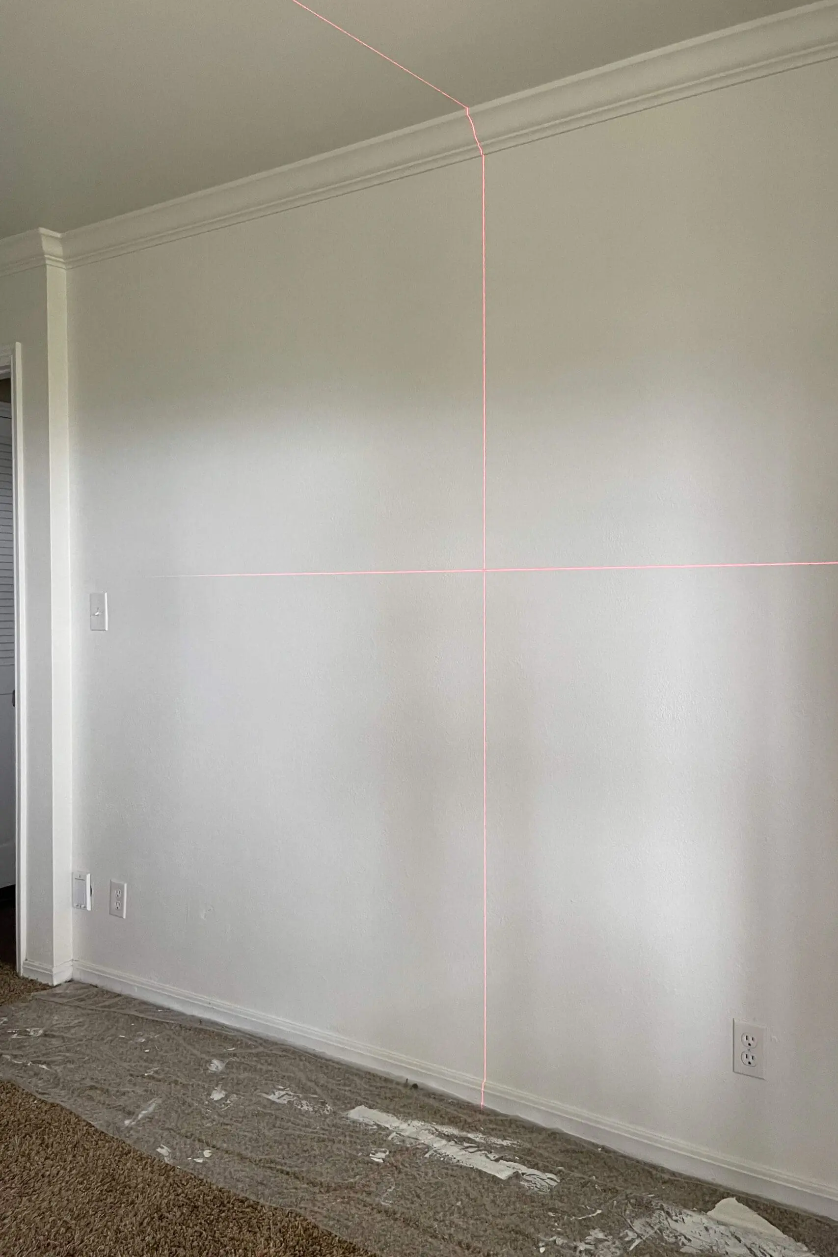 Laser level projecting vertical and horizontal guide lines onto a freshly painted wall to plan a beginner board and batten layout.