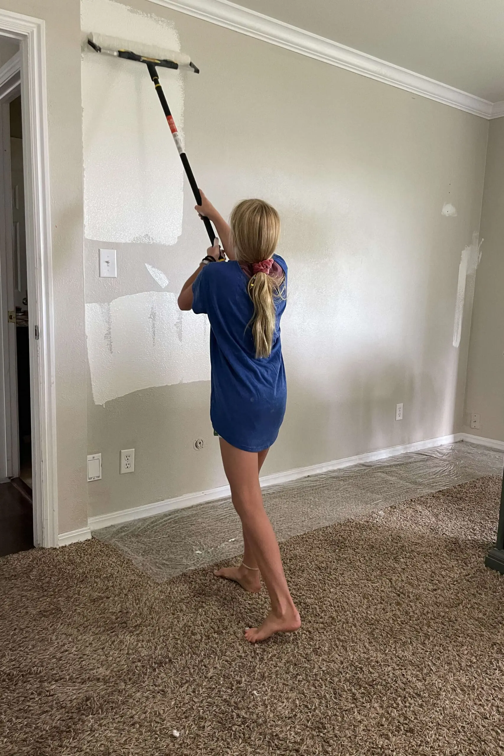 Young girl using a paint roller to cover a beige wall with white paint.