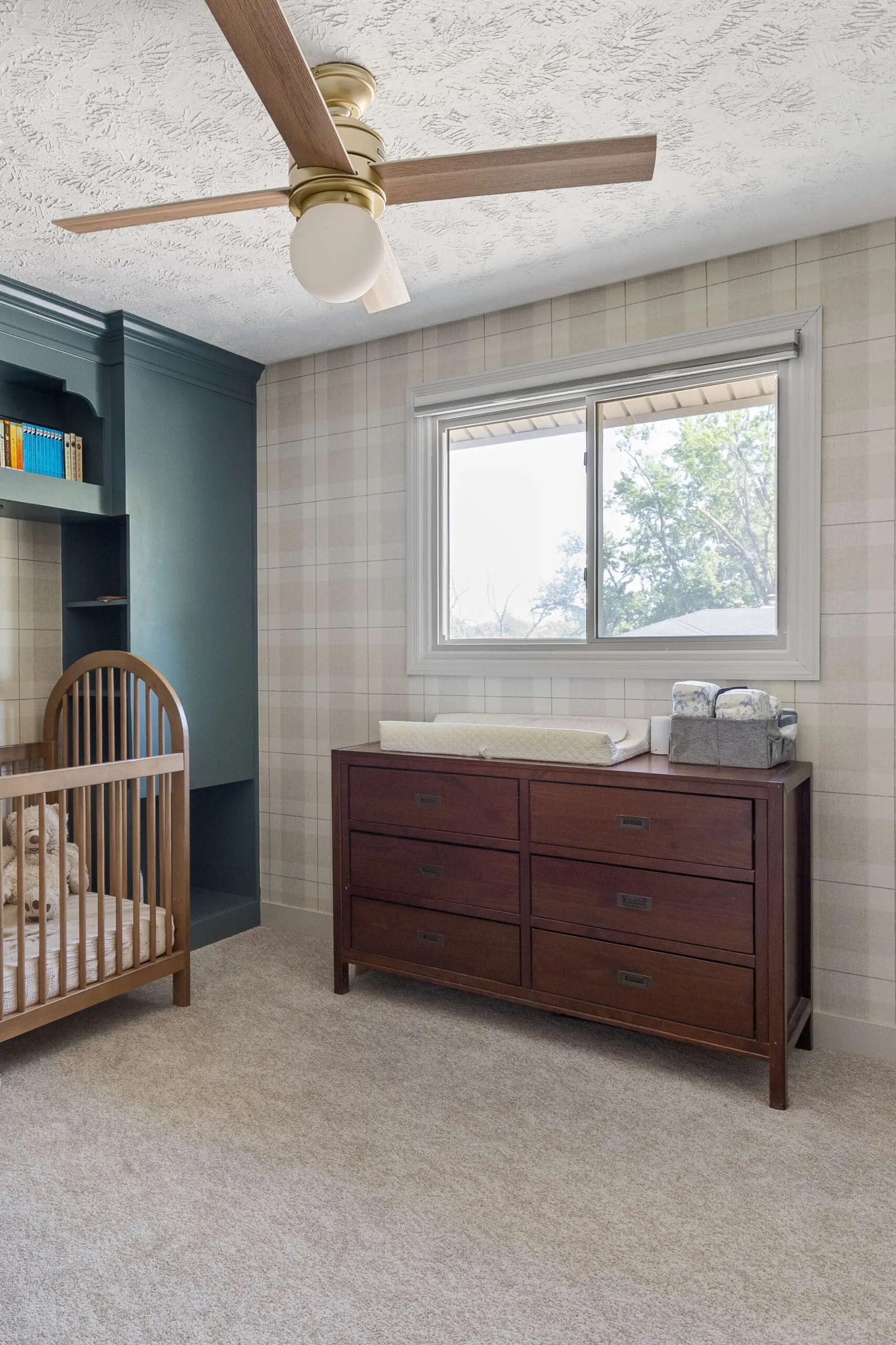 Cozy nursery featuring neutral plaid wallpaper, a wooden crib with stuffed animals, a dark wood dresser with a changing pad and diaper caddy, and built-in sage green shelving next to the crib.