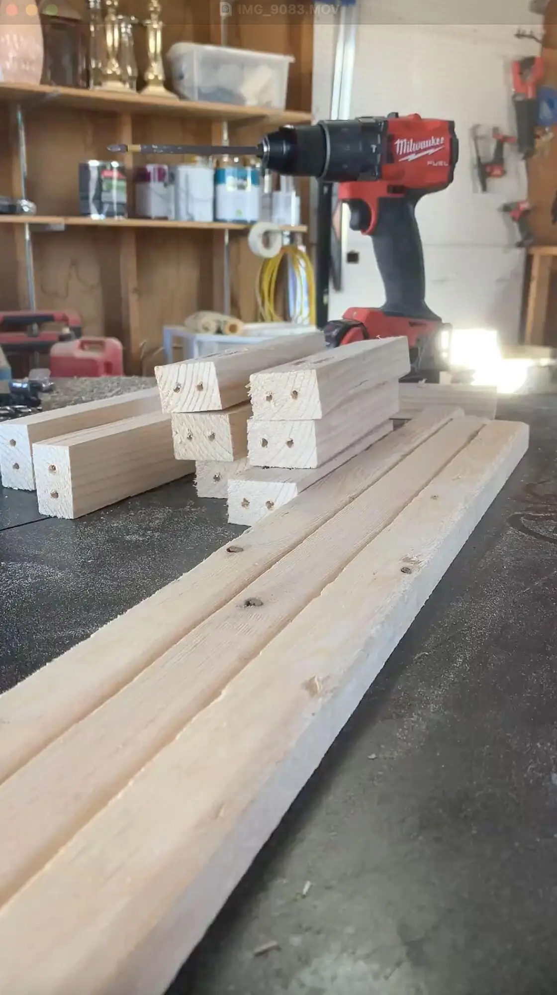 Freshly cut wood boards with pocket holes drilled on the ends, staged on a workbench next to a red power drill in a garage workshop setup.