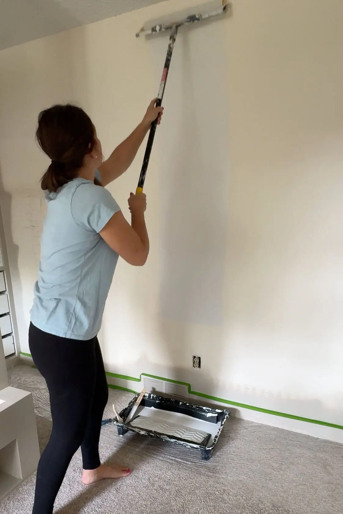 Woman using a paint roller with an extension pole to apply white primer to a beige wall.