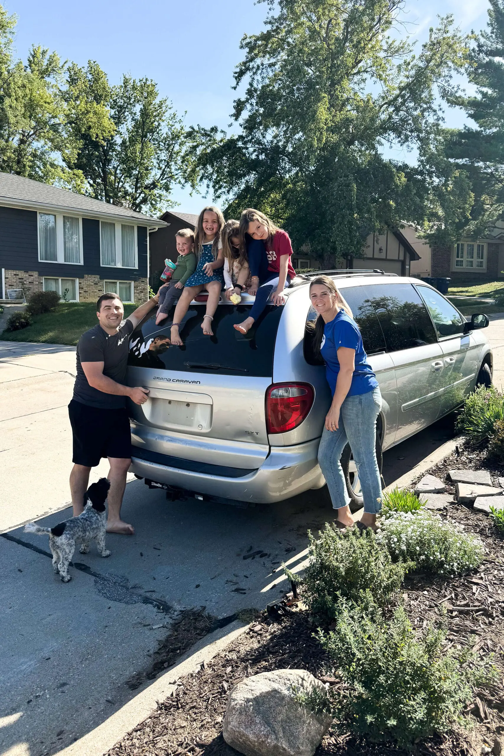Family getting ready for a road trip, with kids smiling and sitting on top of a silver minivan while parents and their dog stand nearby on a sunny day.