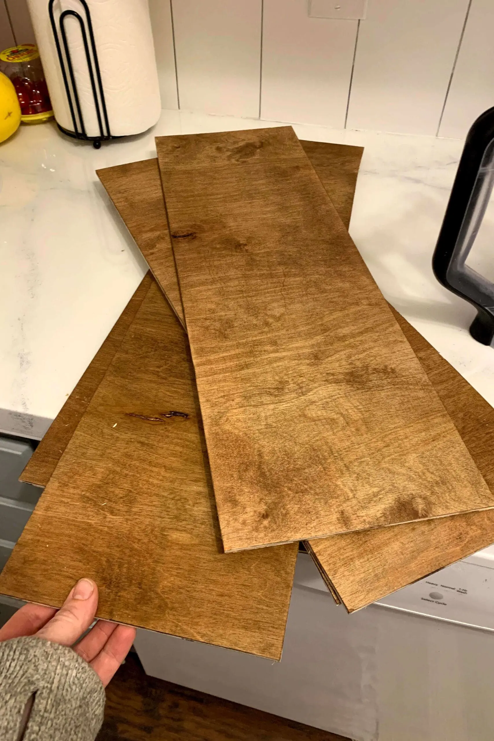 Freshly cut and stained plywood boards laid out on a white kitchen counter, prepped as outer pieces for custom DIY floating shelves.