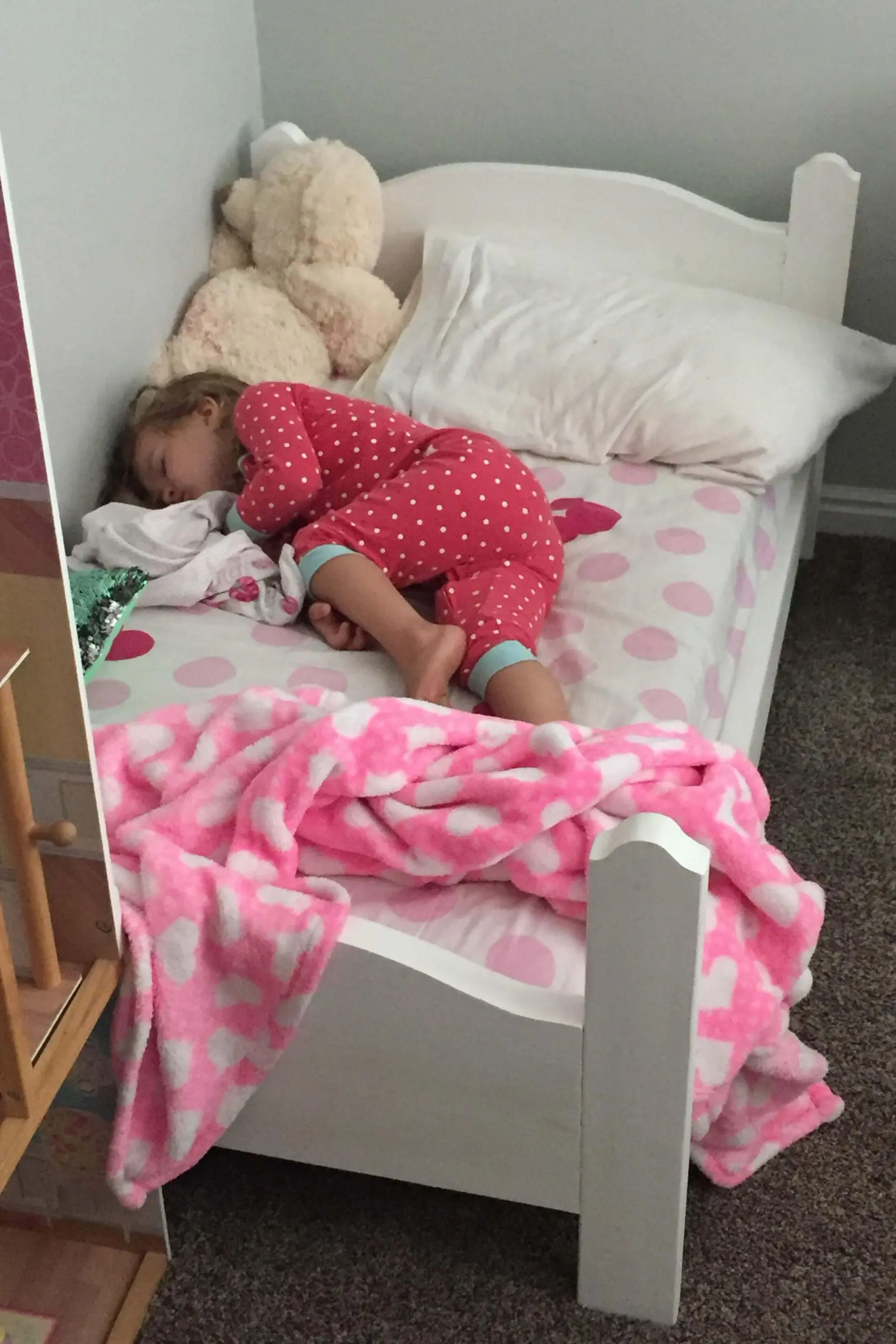 Toddler peacefully asleep in a handmade wooden bed with white paint and pink polka dot bedding, cozy under a soft pink blanket.