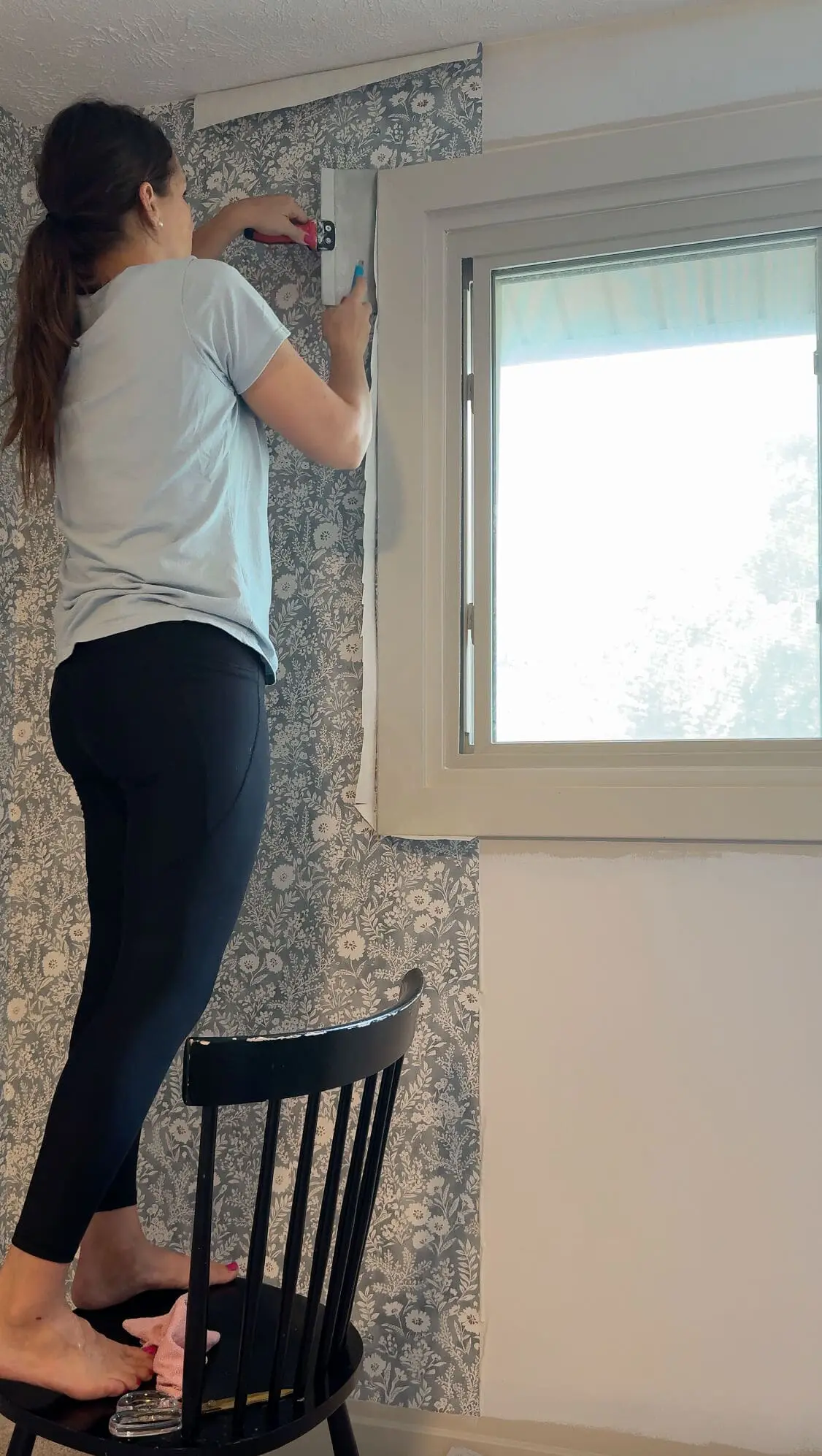 Standing on a chair, woman trims wallpaper along the top corner of a window using a smoothing tool and razor blade.