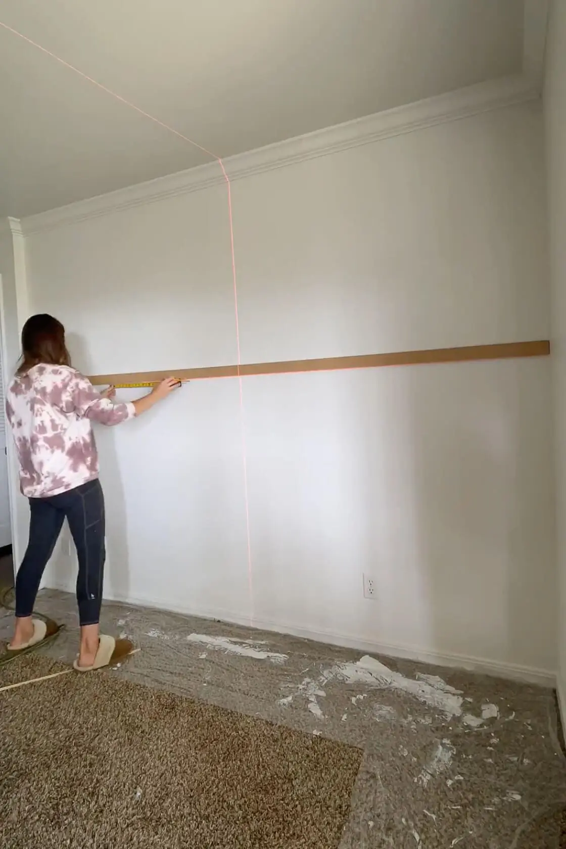 Woman measuring and marking a horizontal trim piece across a painted wall using a laser level to prep for more trim.