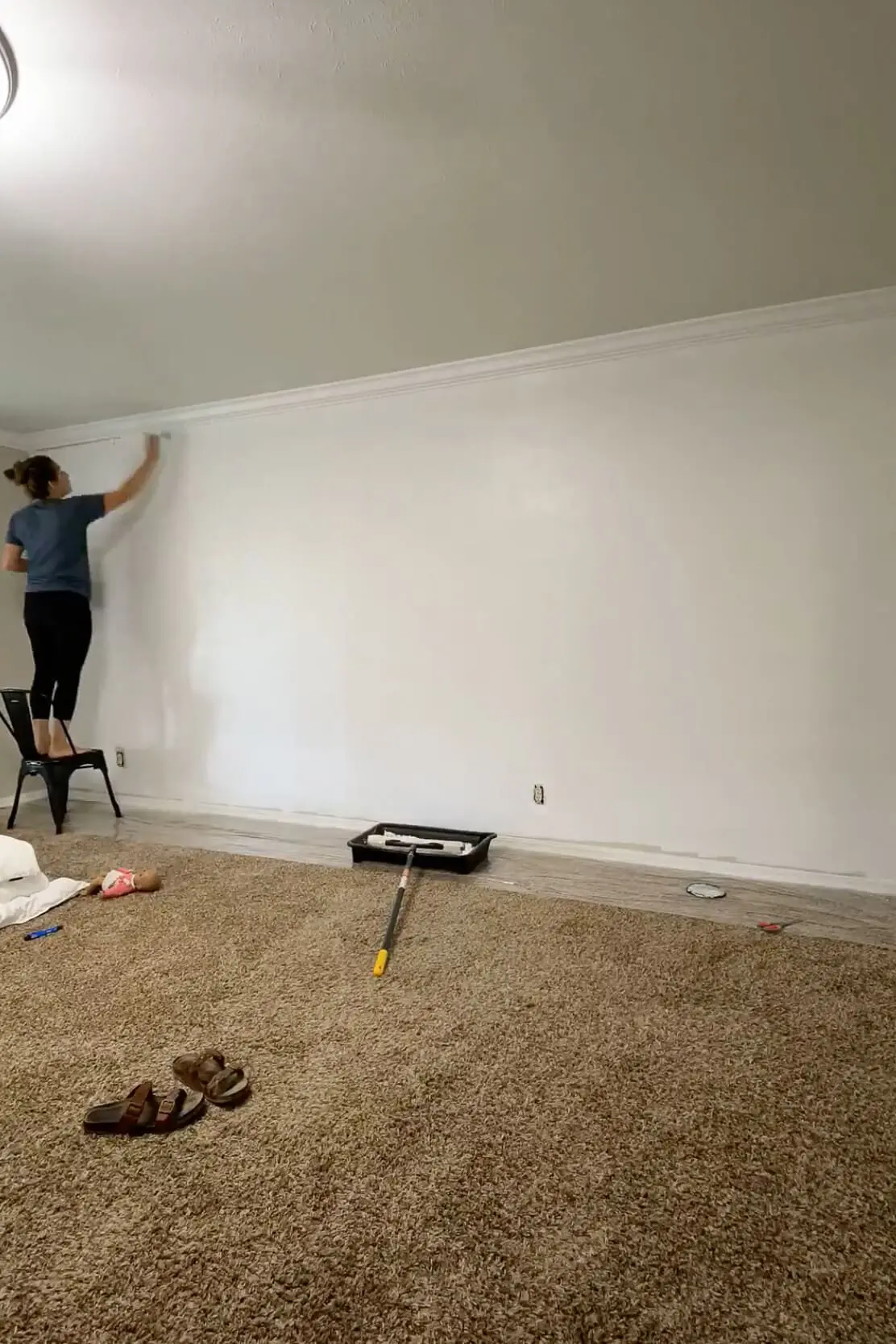 Woman using a roller to paint a wall white while standing on a chair