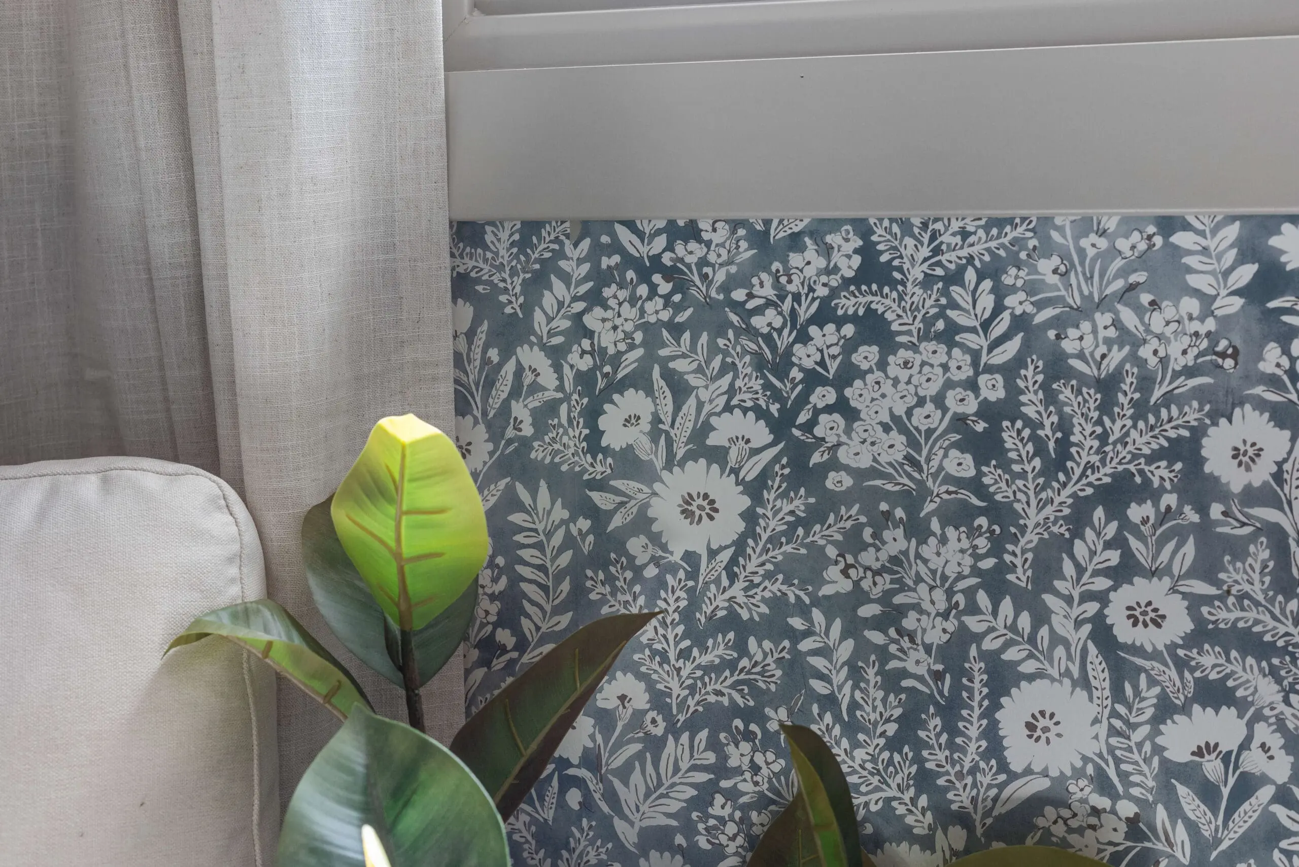 Detailed view of floral wallpaper beneath a window, next to linen curtains and a potted rubber plant.