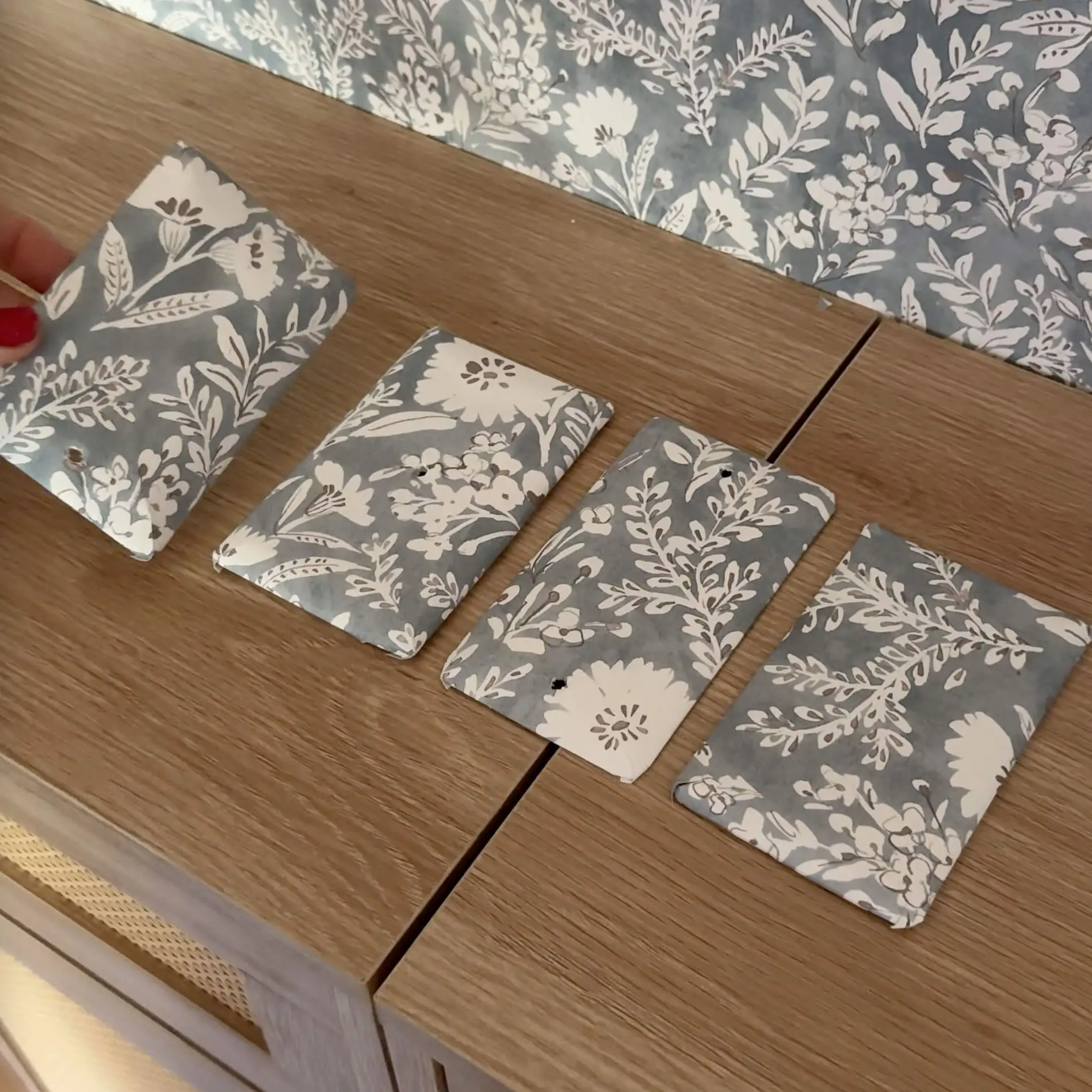 Four outlet and switch plate covers wrapped in floral wallpaper, laid out on a wood cabinet to match the wallpapered wall behind them.