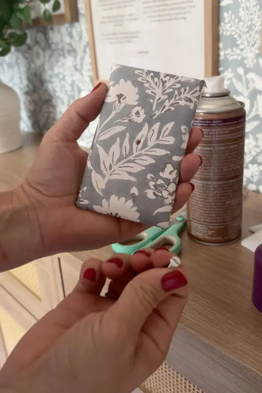 A finished wallpaper-covered switch plate held in hand, showing how the floral pattern wraps neatly around the edges for a polished finish.