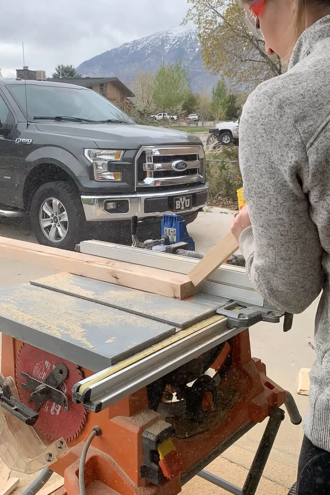 Cutting an angled wood leg on a table saw during a DIY coffee table build, with safety glasses on and a mountain view in the background.