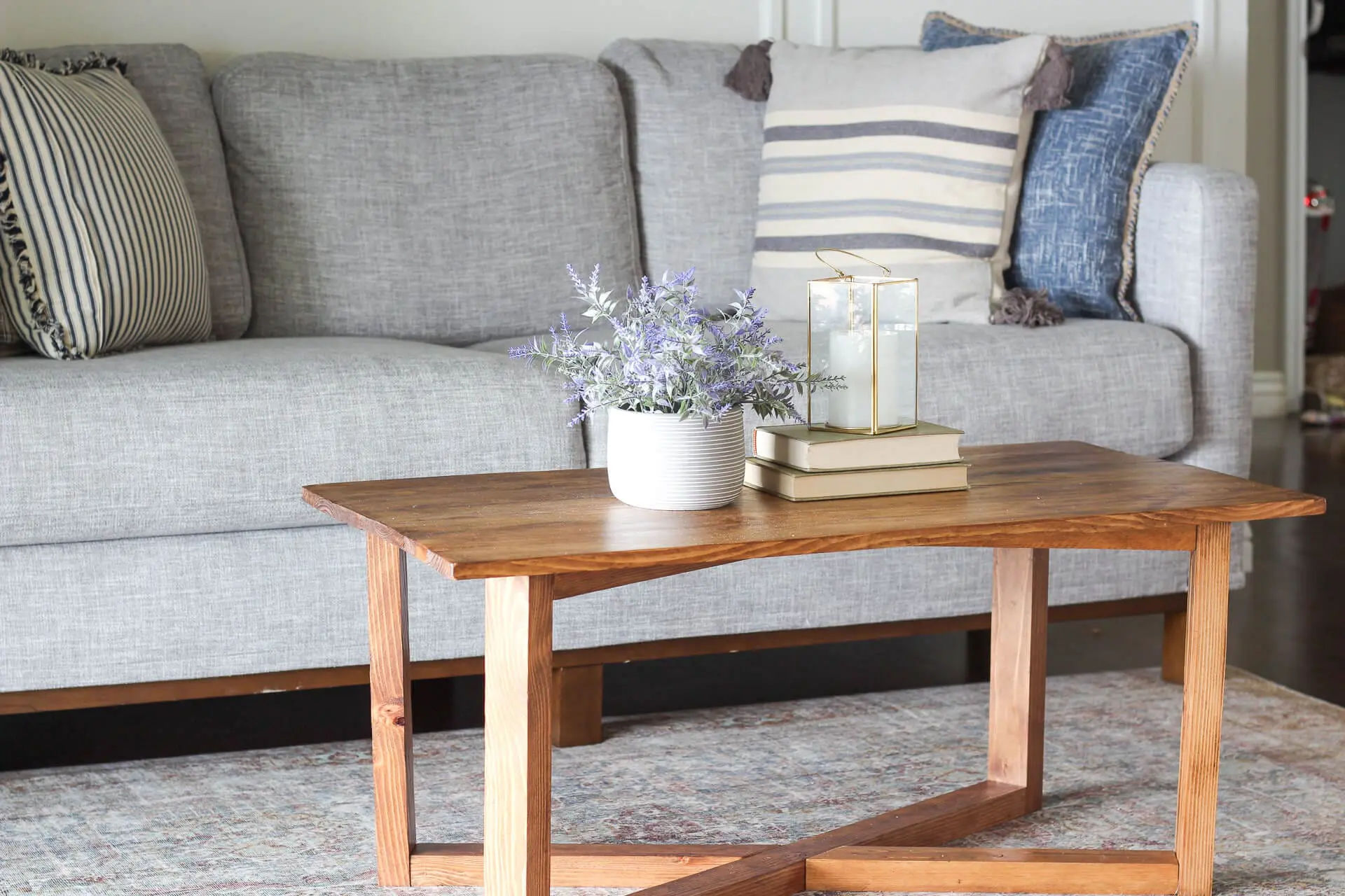 DIY coffee table made from repurposed wood with a natural finish and geometric base, styled in front of a gray couch with striped and textured pillows.