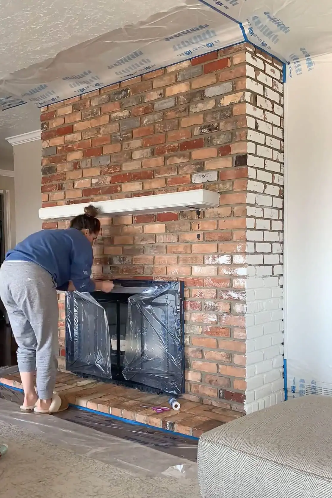 DIY home project in progress with a woman applying plastic masking film along the base of a brick fireplace to prep for an update.