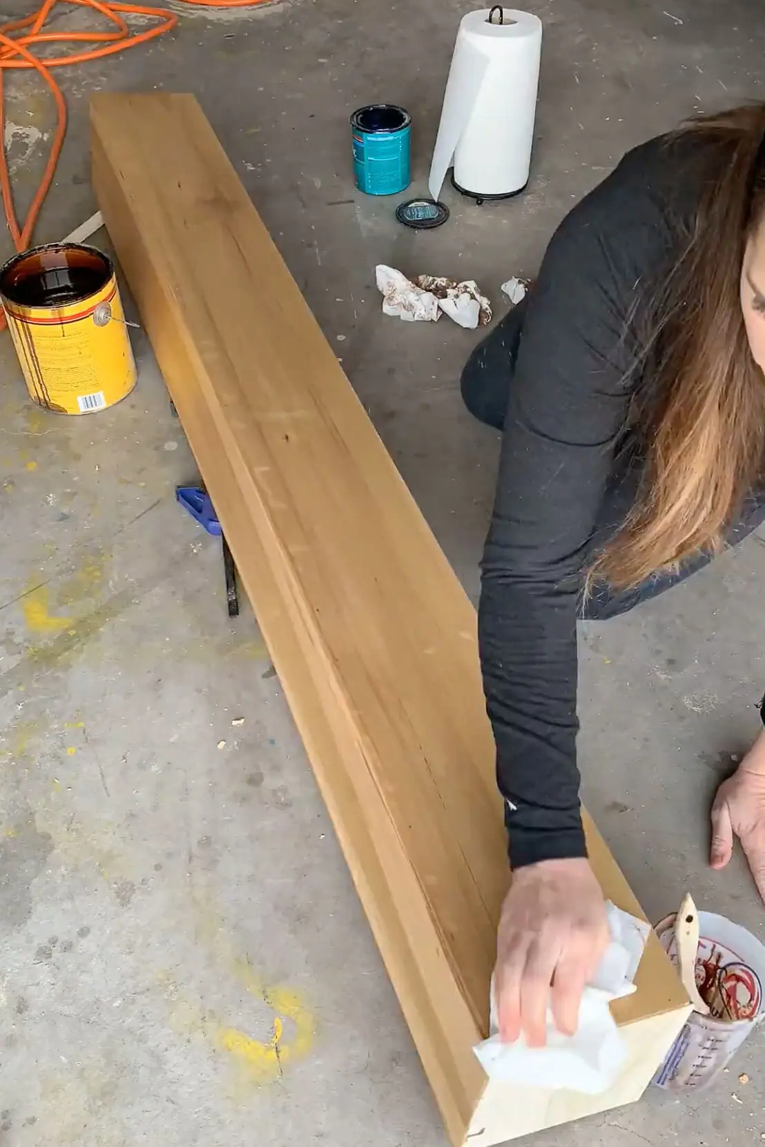 Close-up of a DIY wood mantel being stained by hand in a garage, with cans of stain and supplies visible nearby.