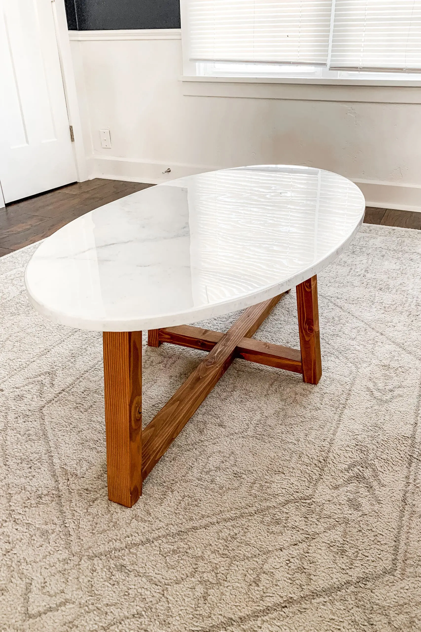 Oval marble-top coffee table with a stained wood base, placed on a neutral area rug in a bright and cozy room.