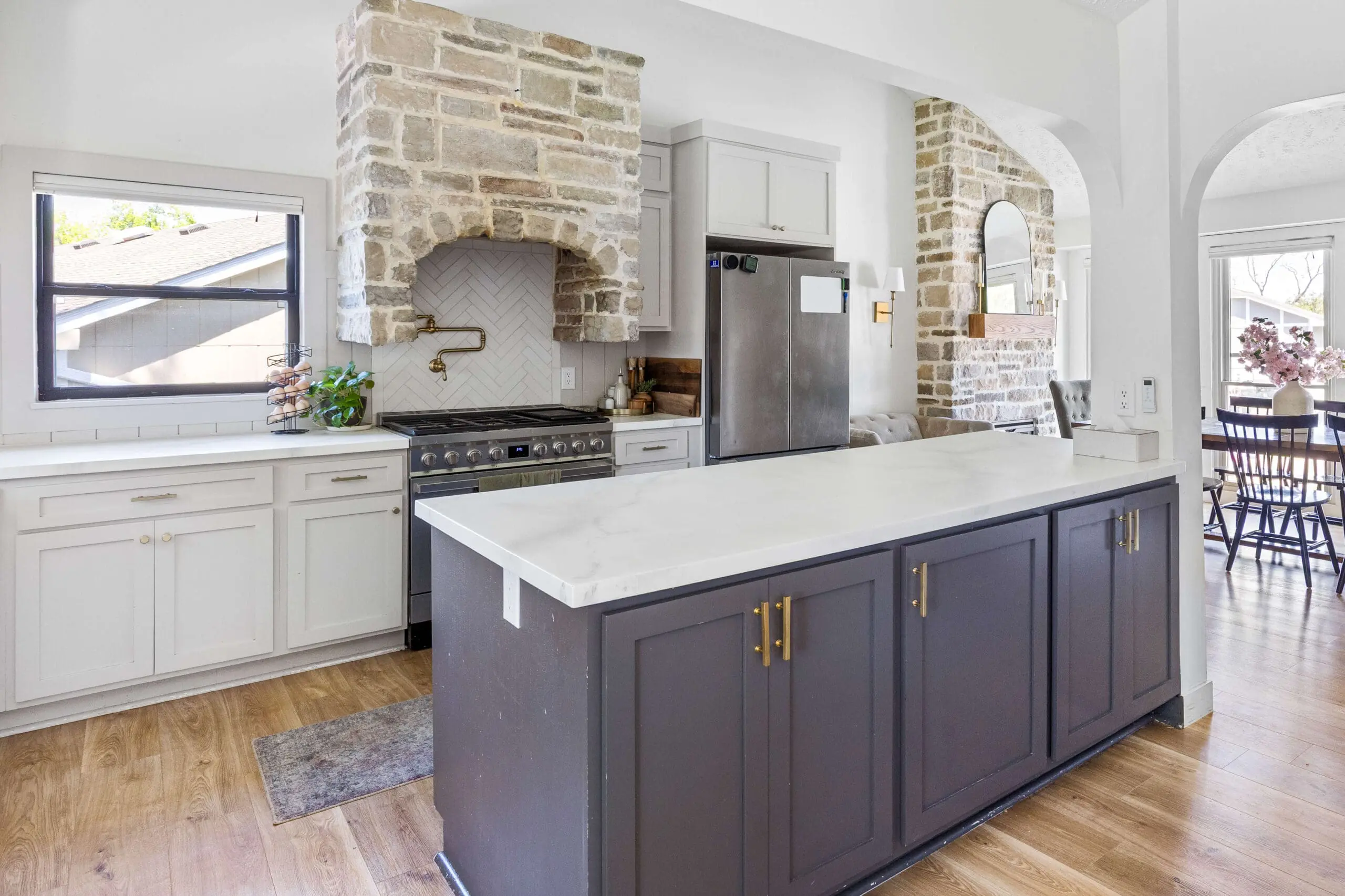 Wide view of a modern kitchen with epoxy faux marble countertops, dark island cabinetry, and a stone range hood with herringbone tile backsplash