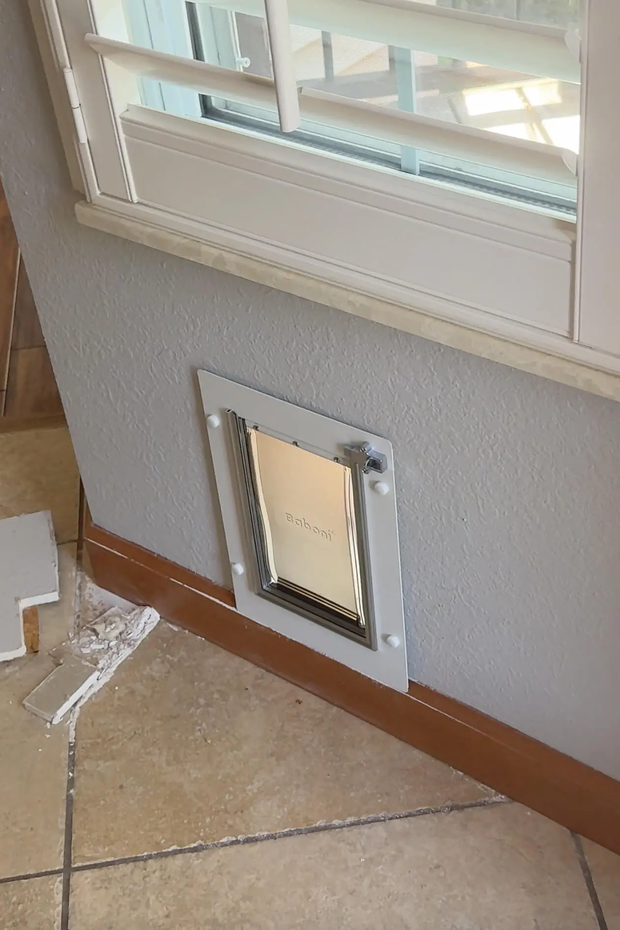 Completed dog door installation on an interior wall beneath a window with plantation shutters and tile flooring.