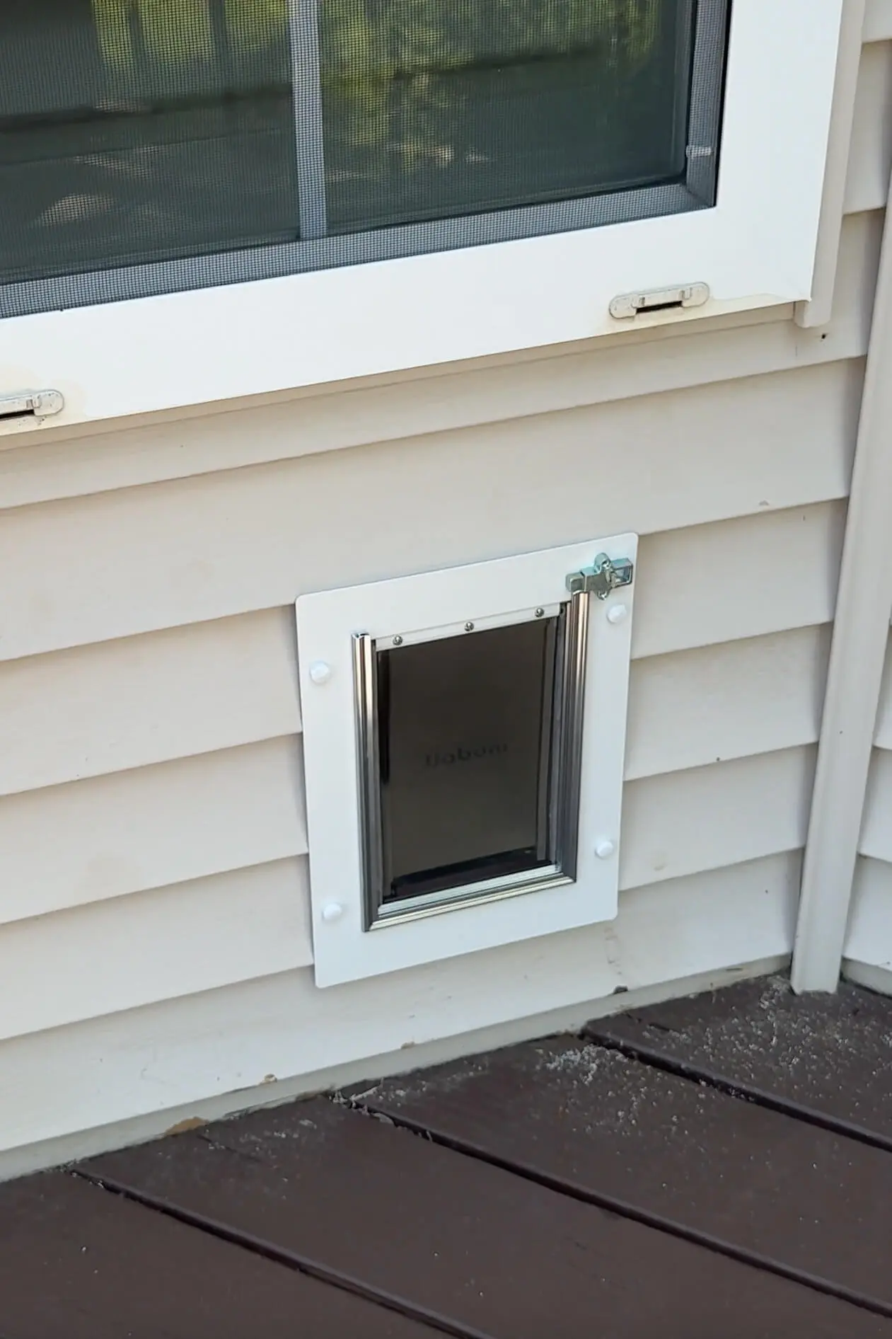 Close-up view of a white-framed dog door installed on tan vinyl siding beneath a window.