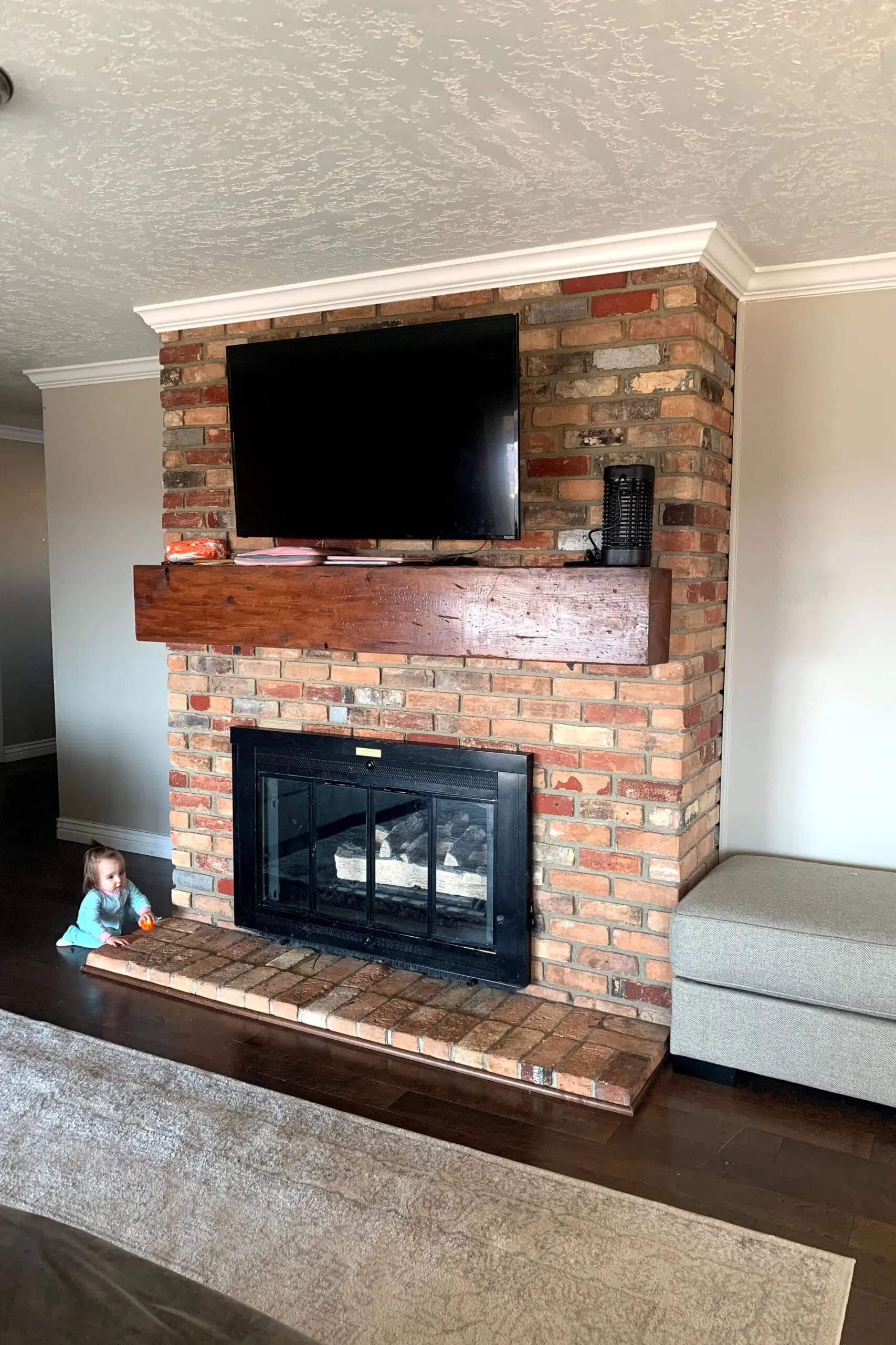 Old, dated, gas insert with rustic red and tan bricks, a dark wood mantel, and a mounted TV above. A toddler sits nearby on the raised hearth.