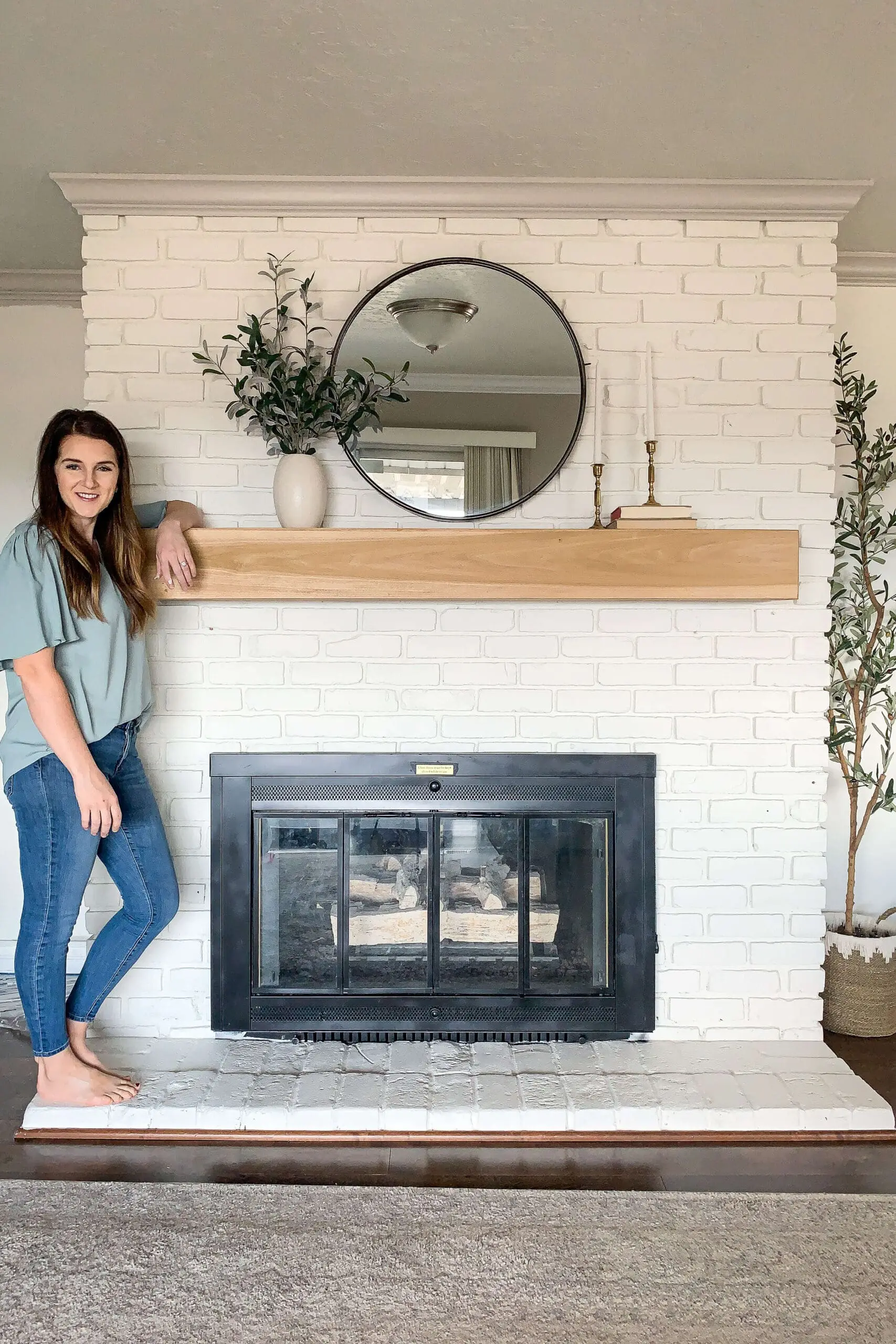 Woman standing beside a white painted brick fireplace with a modern black firebox, round mirror, and natural wood mantel.