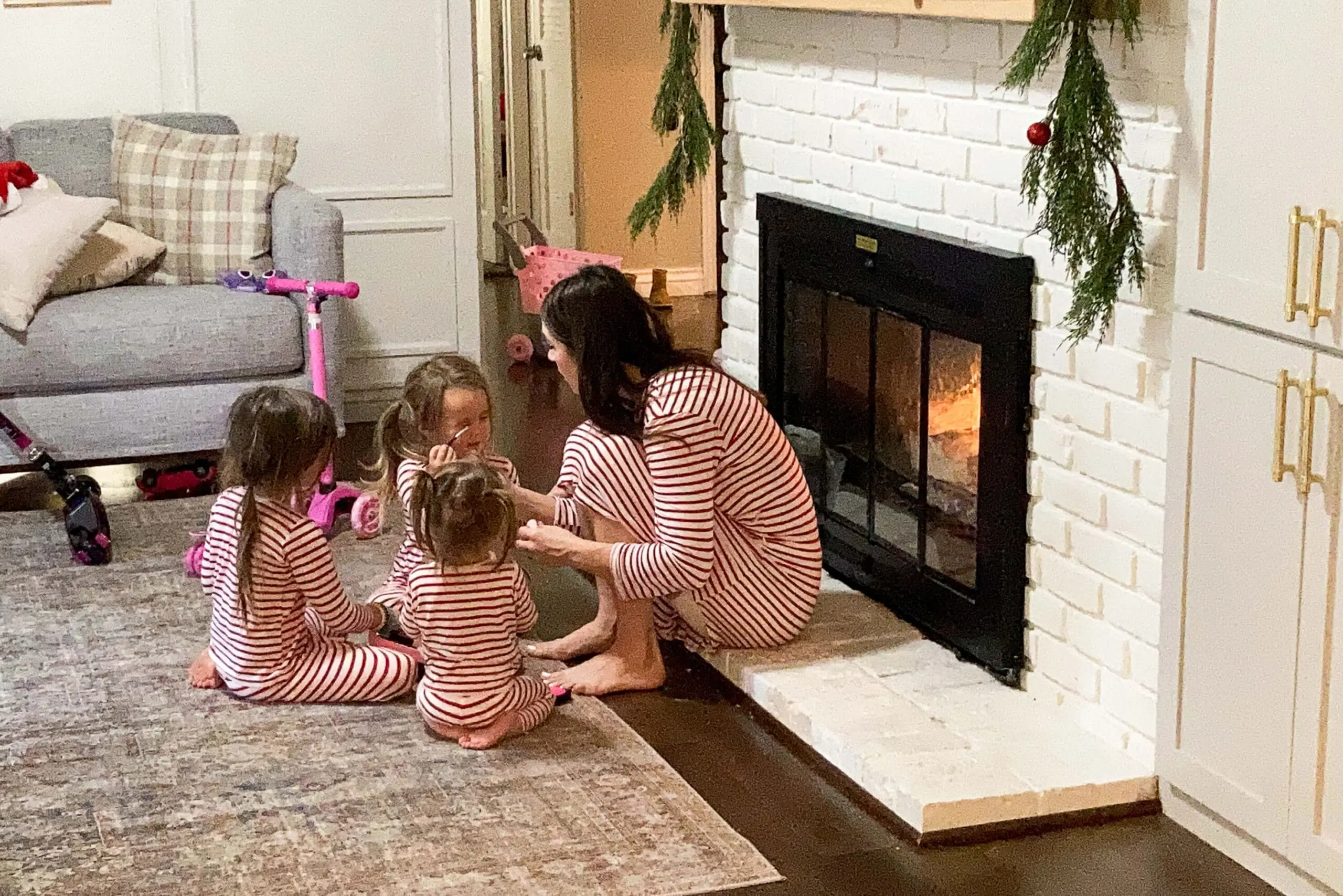 Mom and kids in matching striped pajamas gathered around a cozy fire beside a painted brick fireplace decorated with holiday greenery.
