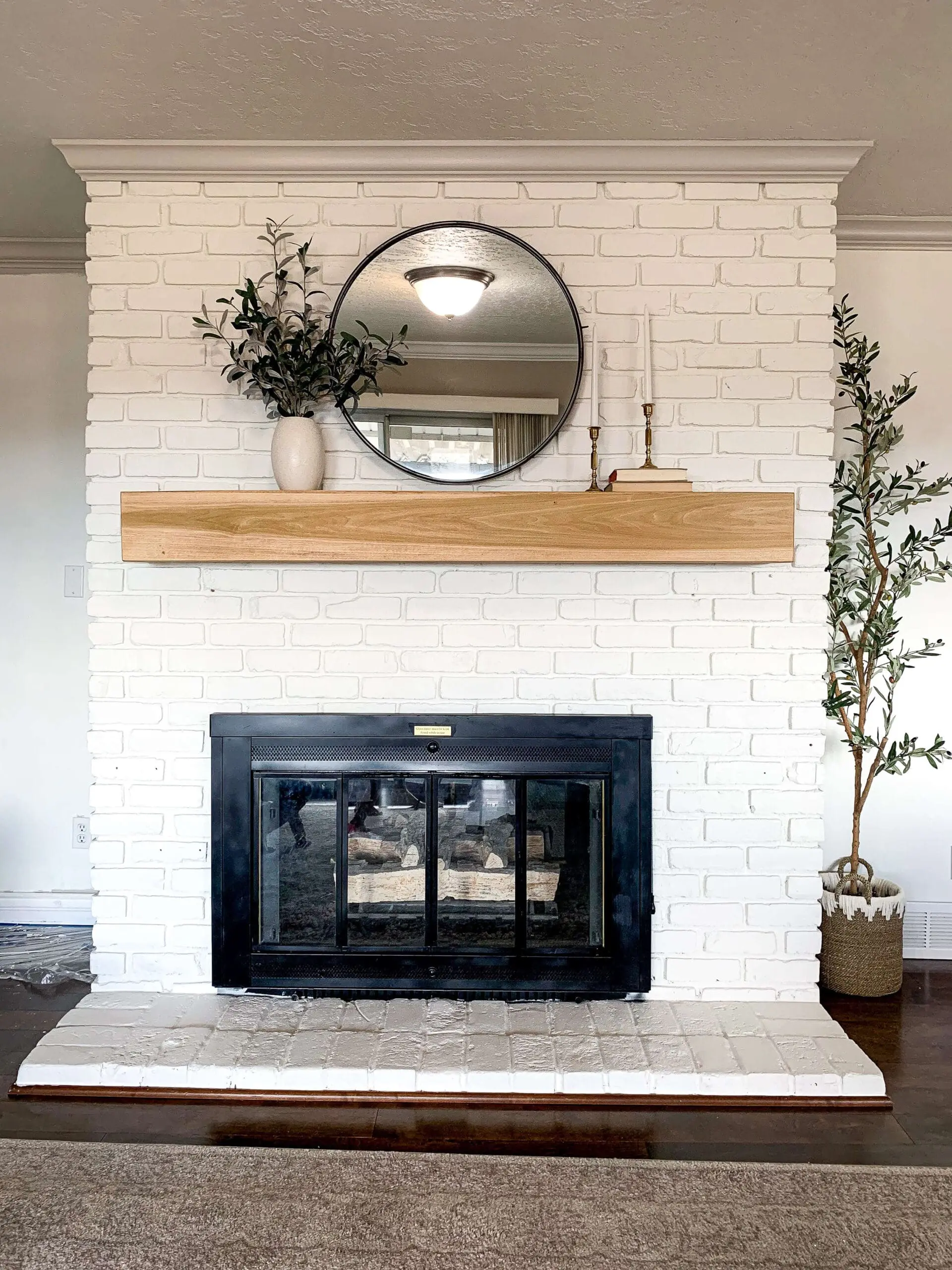 Straight-on view of a white painted brick fireplace with a natural wood mantel, black firebox, and styled with greenery and modern decor.