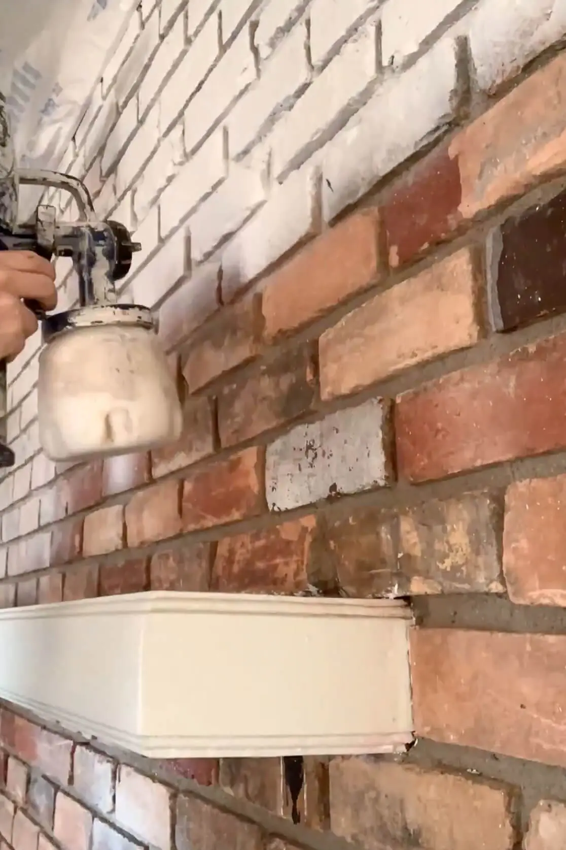 Close-up of a DIYer using a paint sprayer to coat red brick with white paint during a painted brick fireplace transformation.
