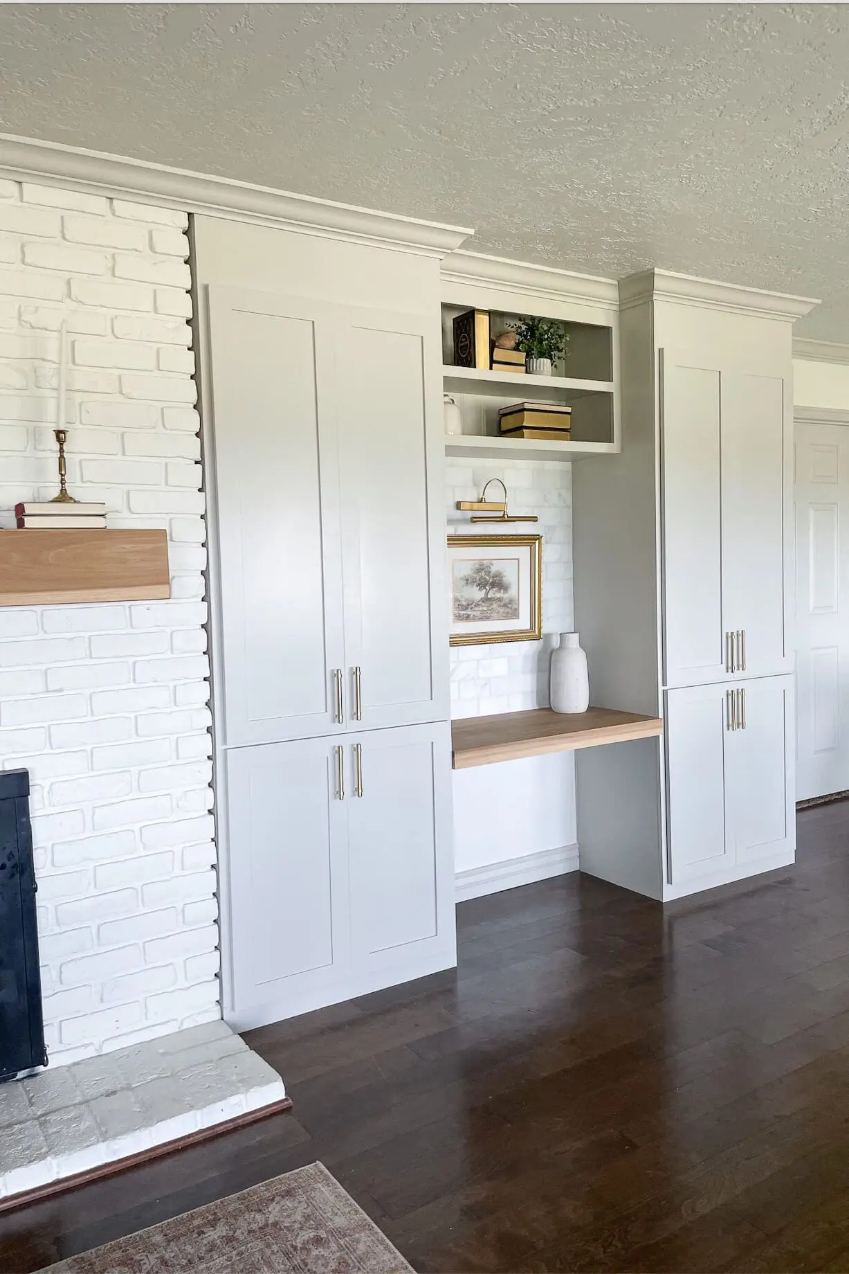 Freshly updated living room with white built-in cabinets and desk space next to a painted brick fireplace with a light wood mantel.