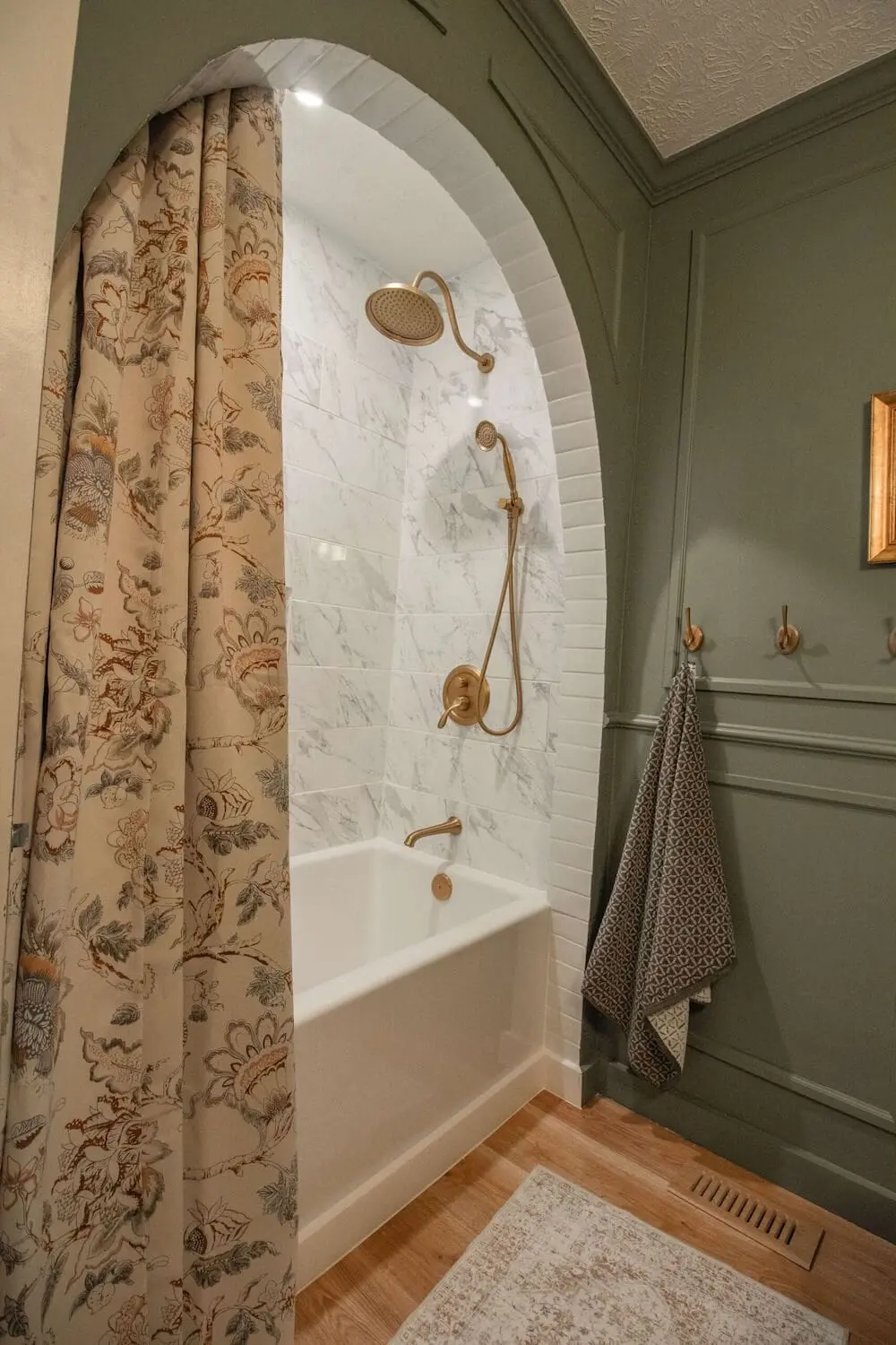 Arched bathtub alcove featuring marble-look tile walls and brushed gold shower fixtures for a high-end, classic bathroom design.