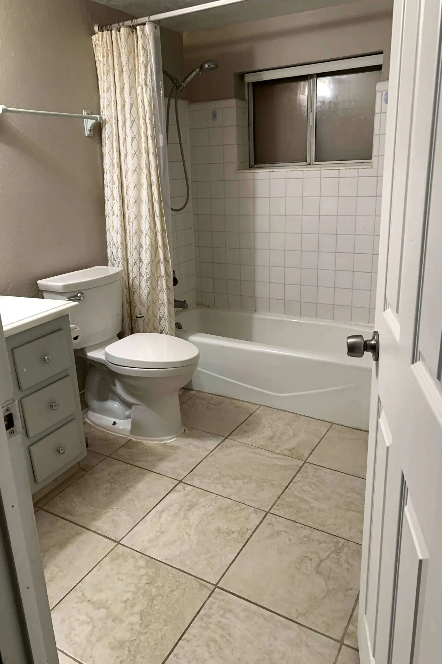 Before photo of the dated wash room, a white toilet, cream vanity, and old shower and small window.