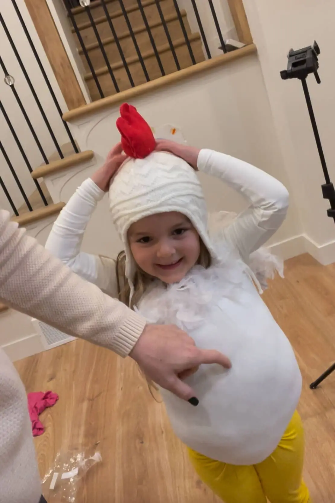 Smiling child wearing a white knit hat with a red felt comb and feathered shirt while trying on a chicken costume in progress.