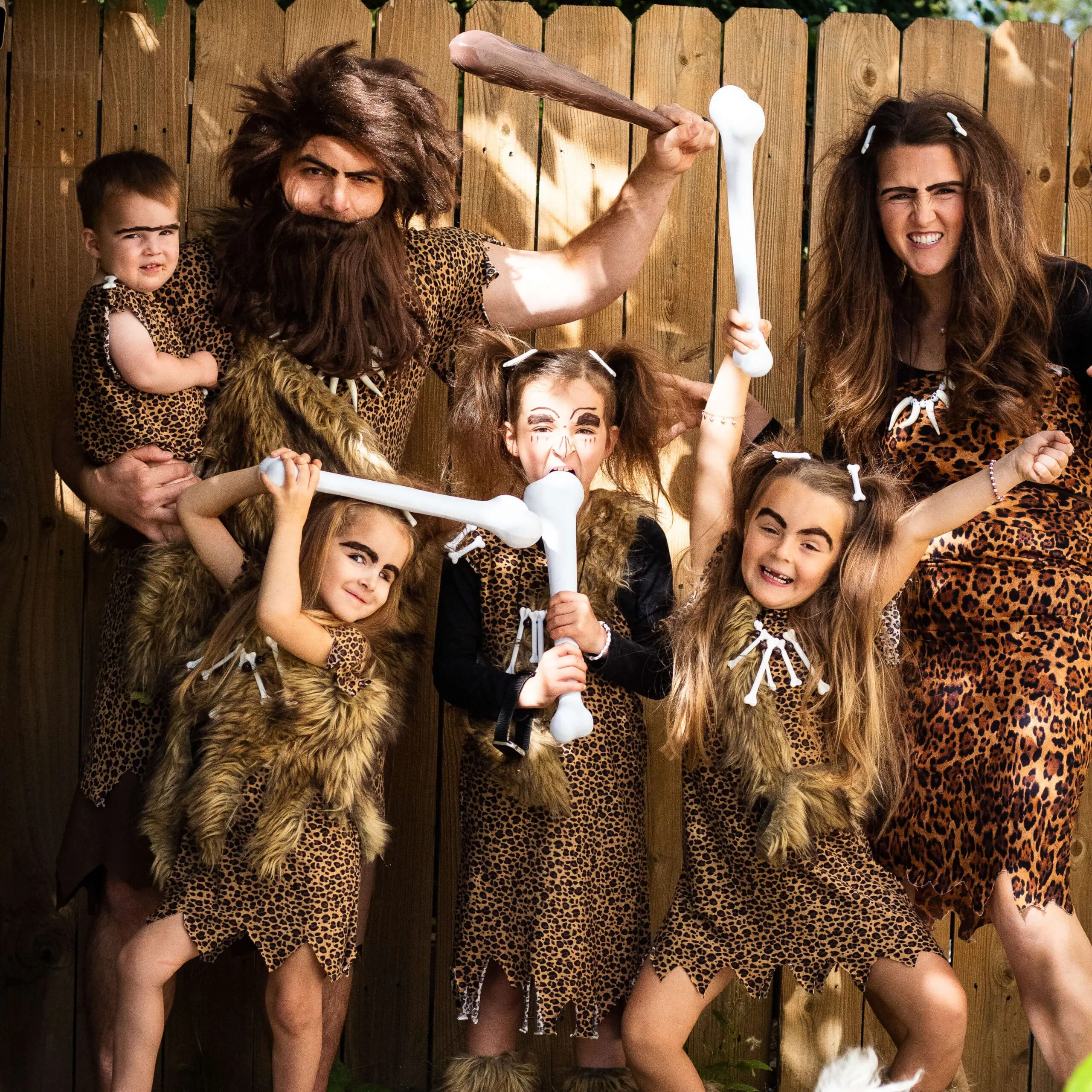 Family costume ideas featuring a fun caveman and cavewoman theme with parents and kids in leopard print outfits, faux fur accessories, and large toy bones.