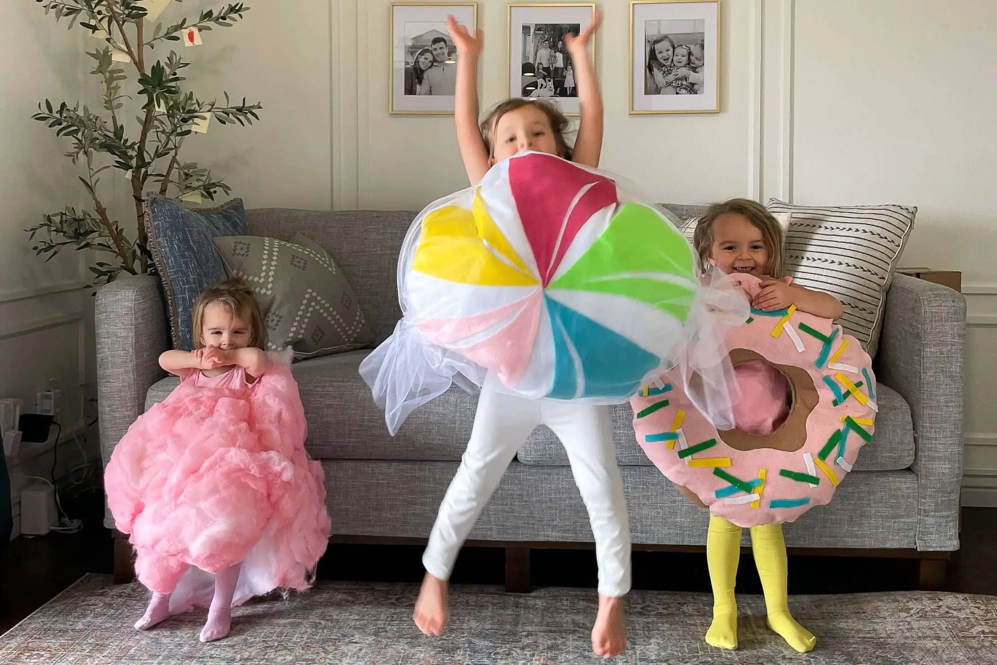 Three kids in candy costumes, including a pink cotton candy outfit, a colorful lollipop costume, and a donut costume with sprinkles, sitting and playing in a living room.