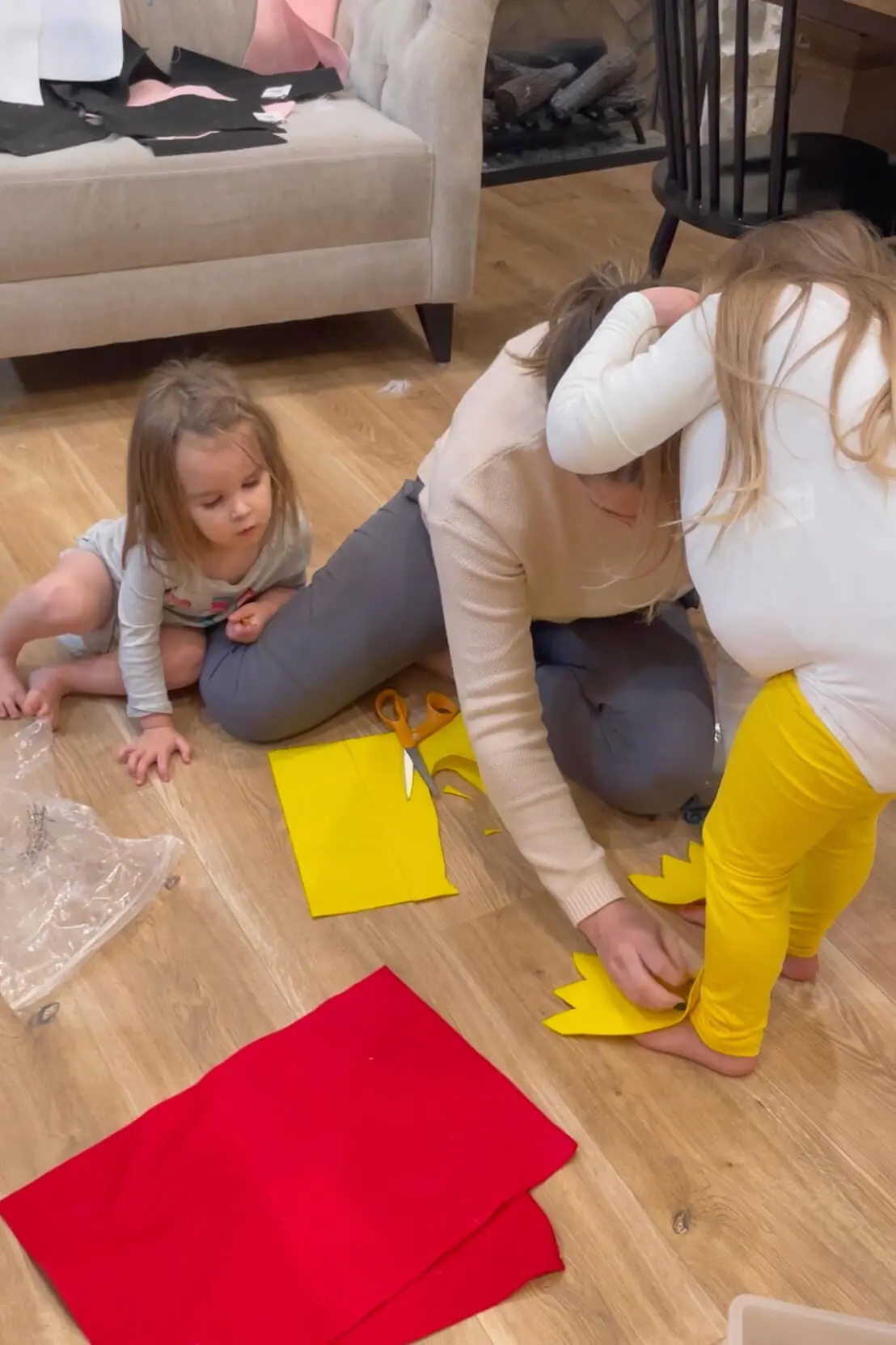Mom helping child fit yellow felt chicken feet while another toddler watches, surrounded by crafting supplies on a wood floor.