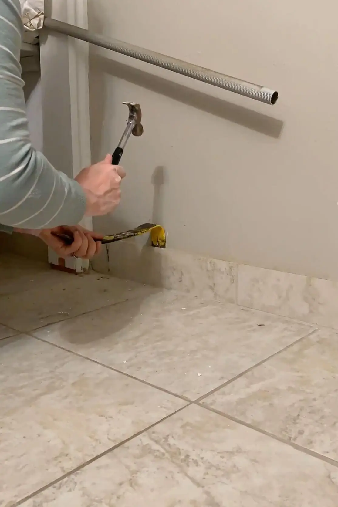 Close-up of baseboard removal during DIY renovation, showing hands using a hammer and pry bar to remove old tile.