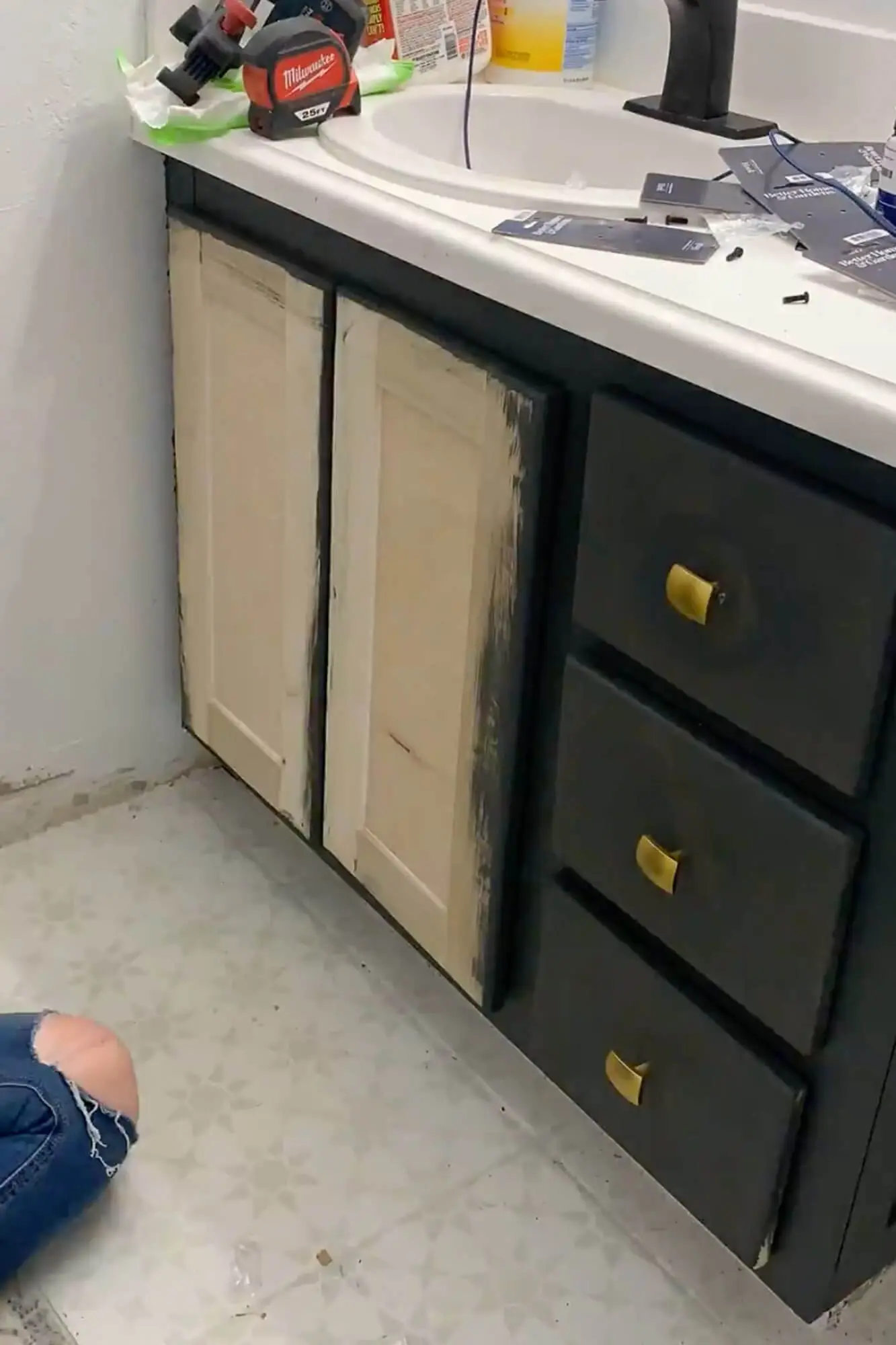 Bathroom vanity mid-makeover with new shaker-style doors in primer and dark painted drawers with gold hardware.