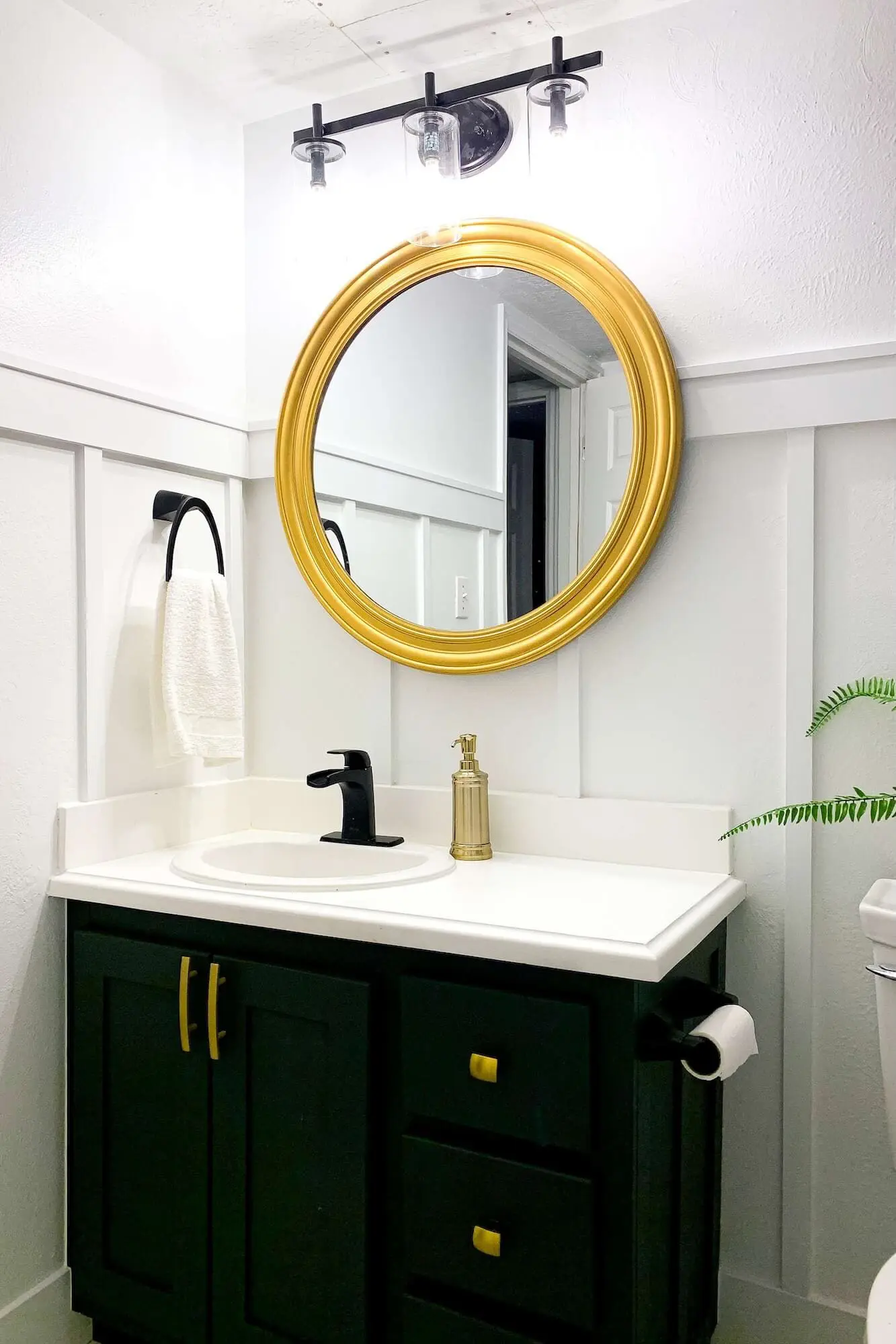 Finished vanity wall with dark green cabinet, gold hardware, gold round mirror, black faucet, and white board and batten backdrop.