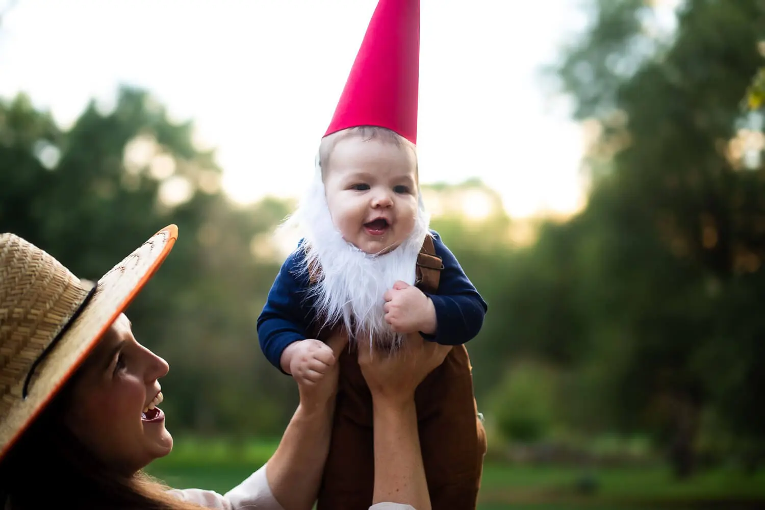 Baby dressed in a handmade garden gnome costume with a red hat, white beard, and brown overalls being lifted up outdoors by a smiling mom wearing a straw gardener hat.