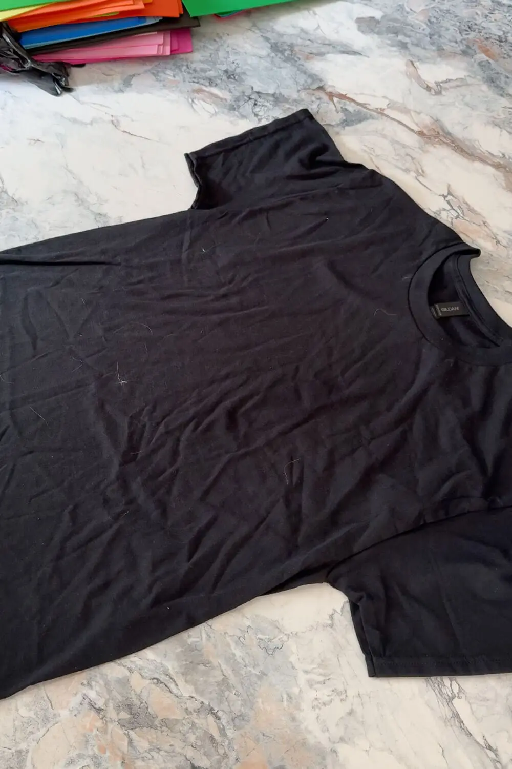 Plain black tee laid flat on a marble countertop with colorful craft paper sheets stacked in the background, ready for a DIY project.