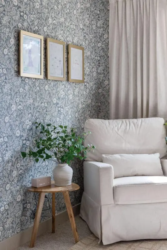 Blue floral wallpaper wall with a cream armchair, linen curtains, three gold frames, and a plant on a wood stool.