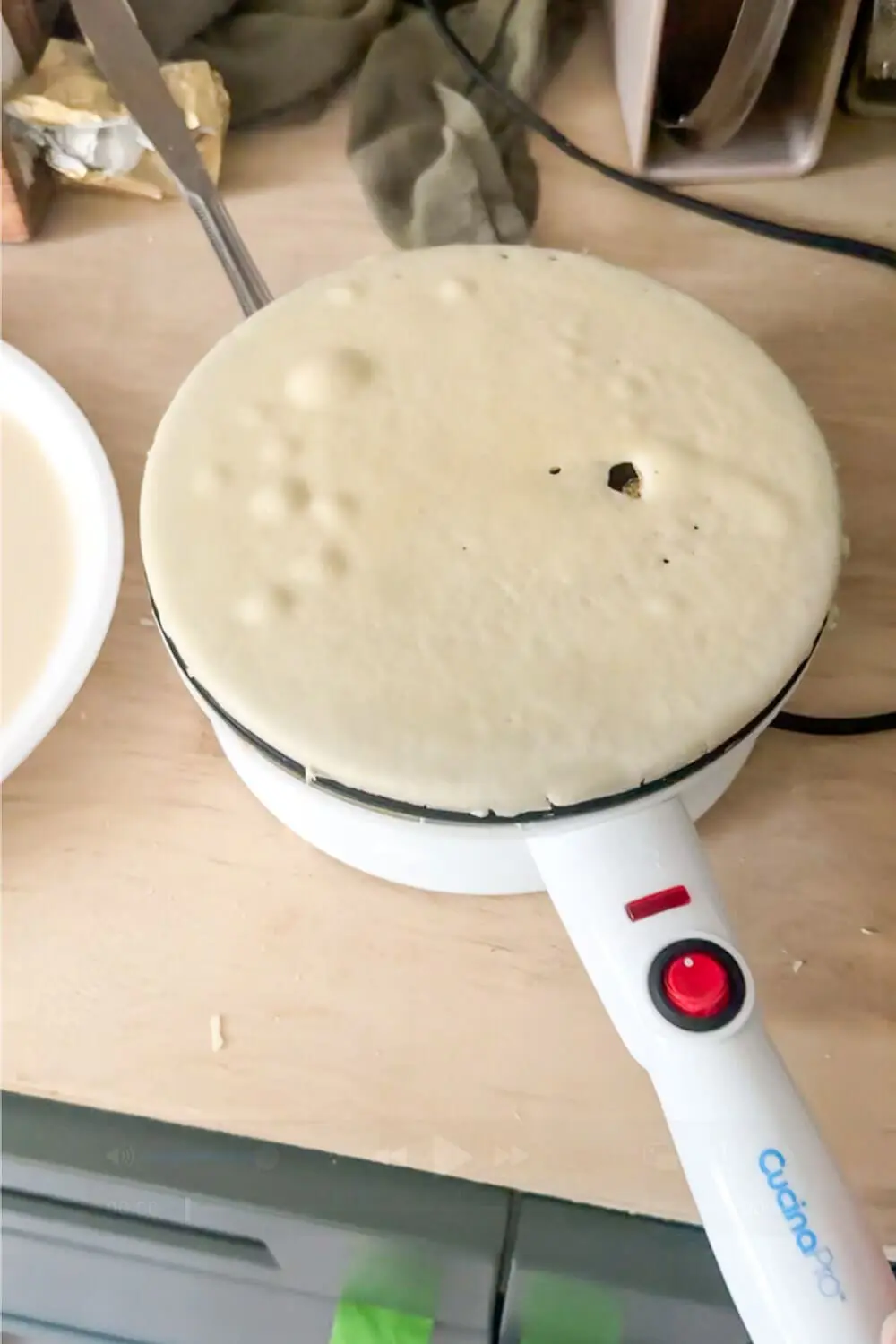 Cordless CucinaPro crepe maker cooking a thin crepe; batter bubbling with a small hole forming and the red power light on.